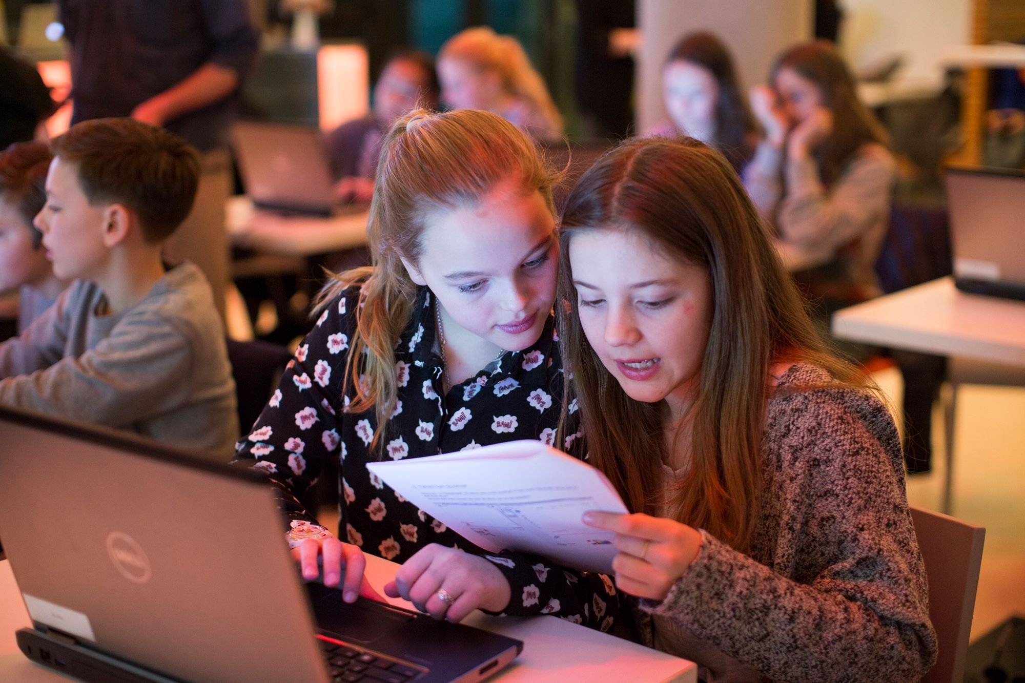 Twee meisjes zitten achter een laptop en kijken op een papier.