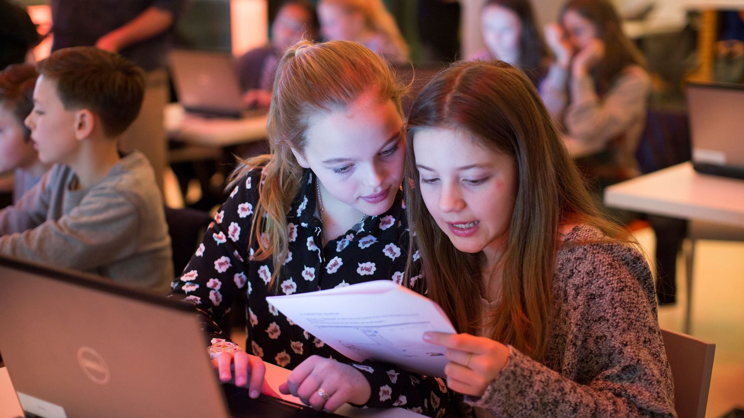 Twee meisjes zitten achter een laptop en kijken op een papier.
