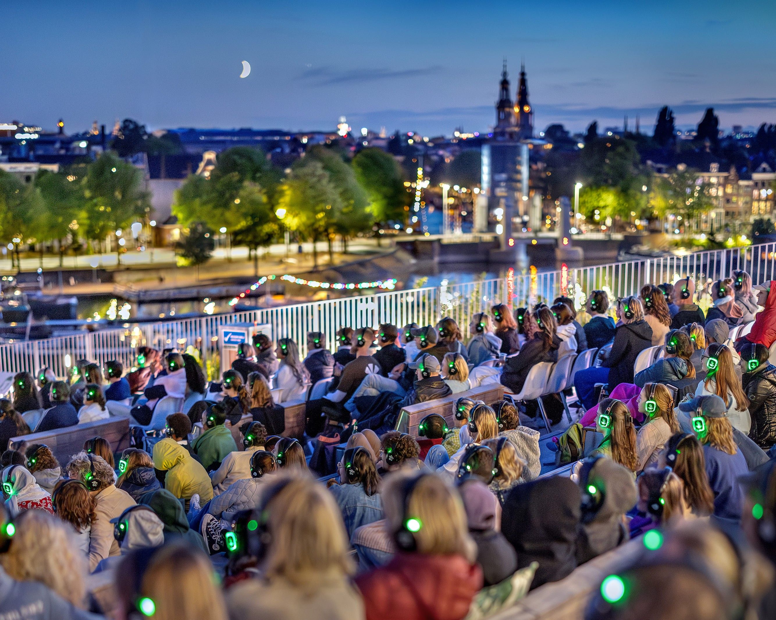 Publiek met koptelefoons op het dak van NEMO en de skyline van Amsterdam op de achtergrond.