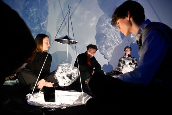 Vier mensen zitten in een ruimte met blauwe projecties op de muren. Tussen hen in staat een kunstinstallatie.