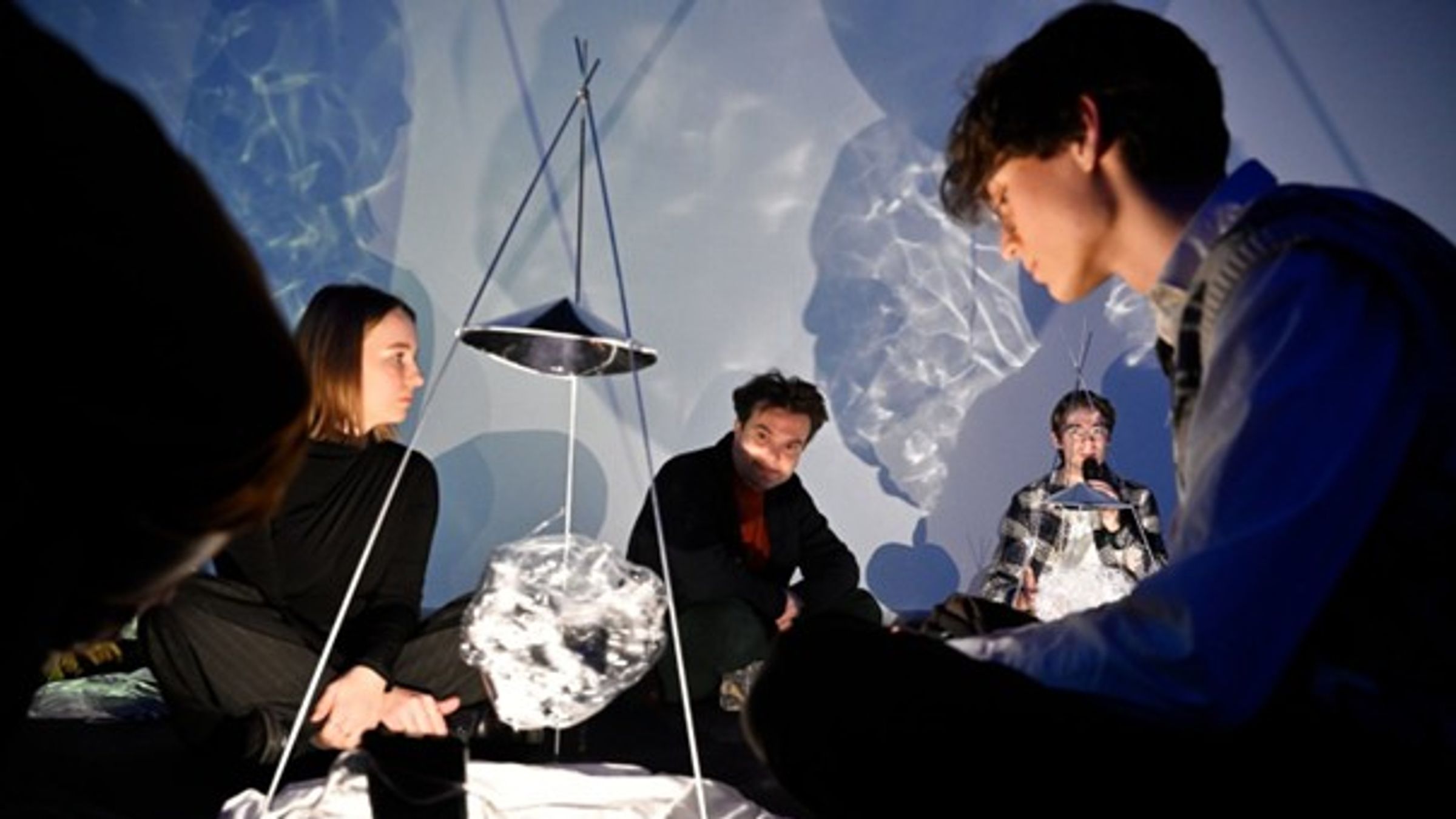 Vier mensen zitten in een ruimte met blauwe projecties op de muren. Tussen hen in staat een kunstinstallatie.