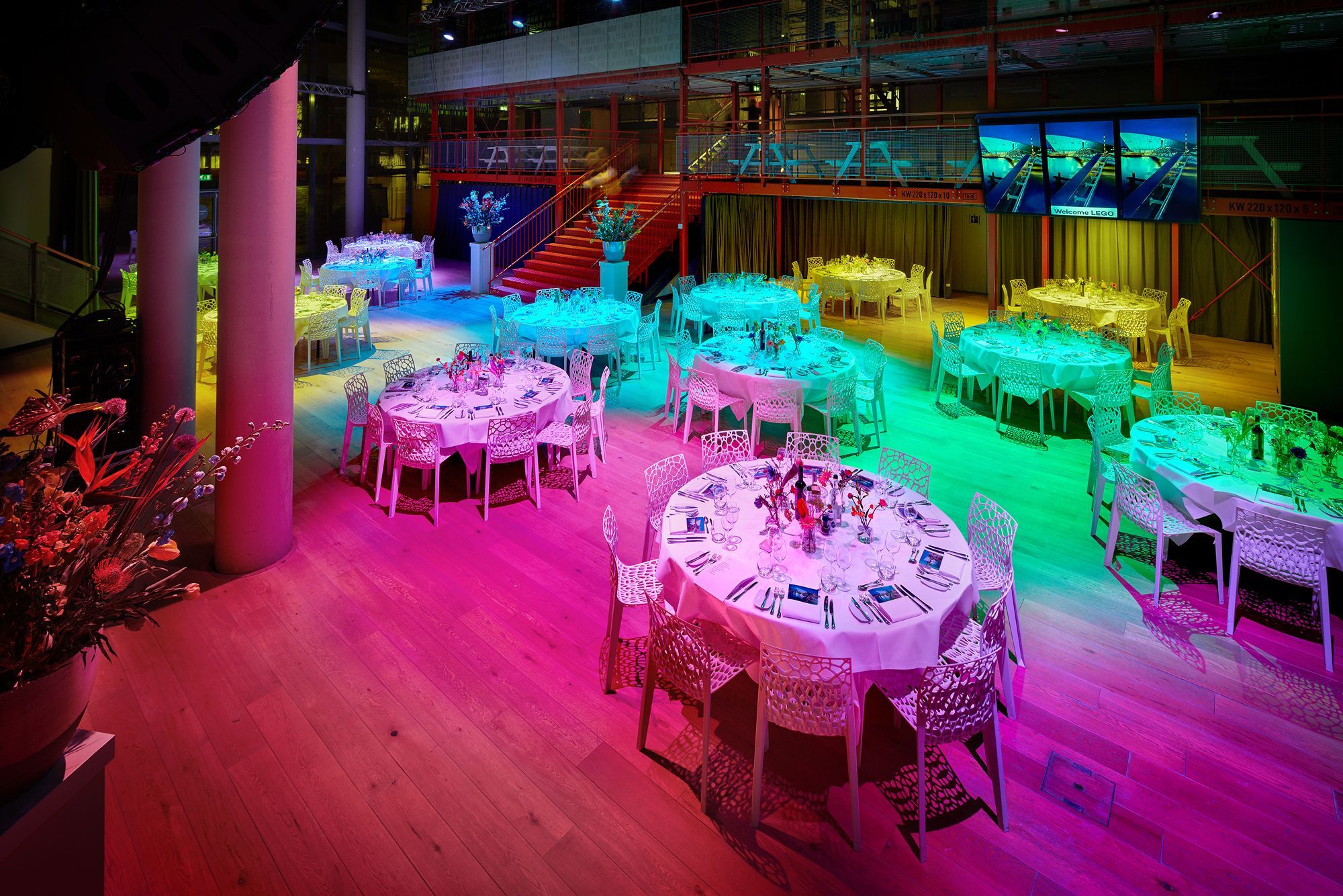 Een gekleurd verlichte zaal met ronde tafels en witte stoelen.