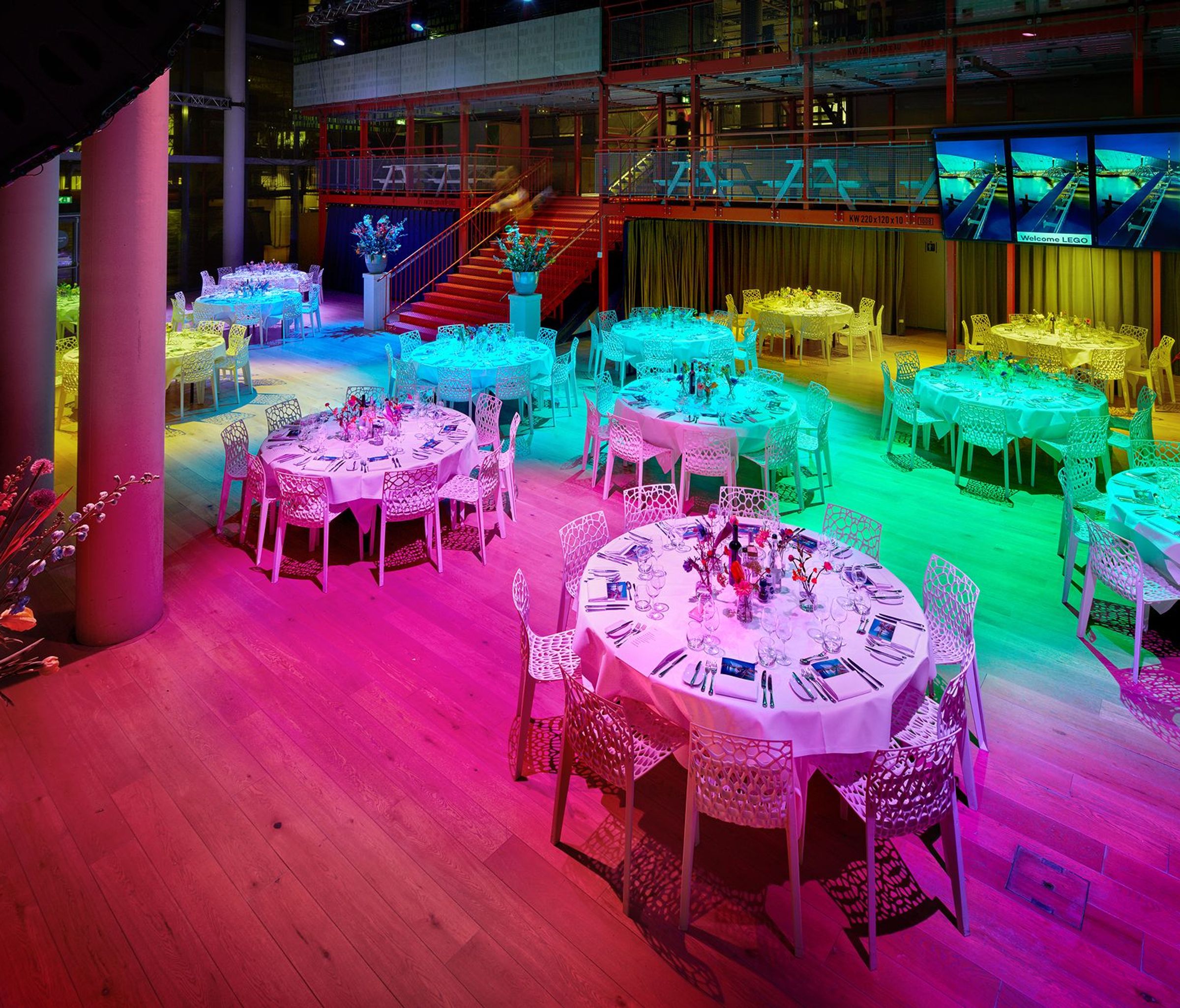 Een gekleurd verlichte zaal met ronde tafels en witte stoelen.
