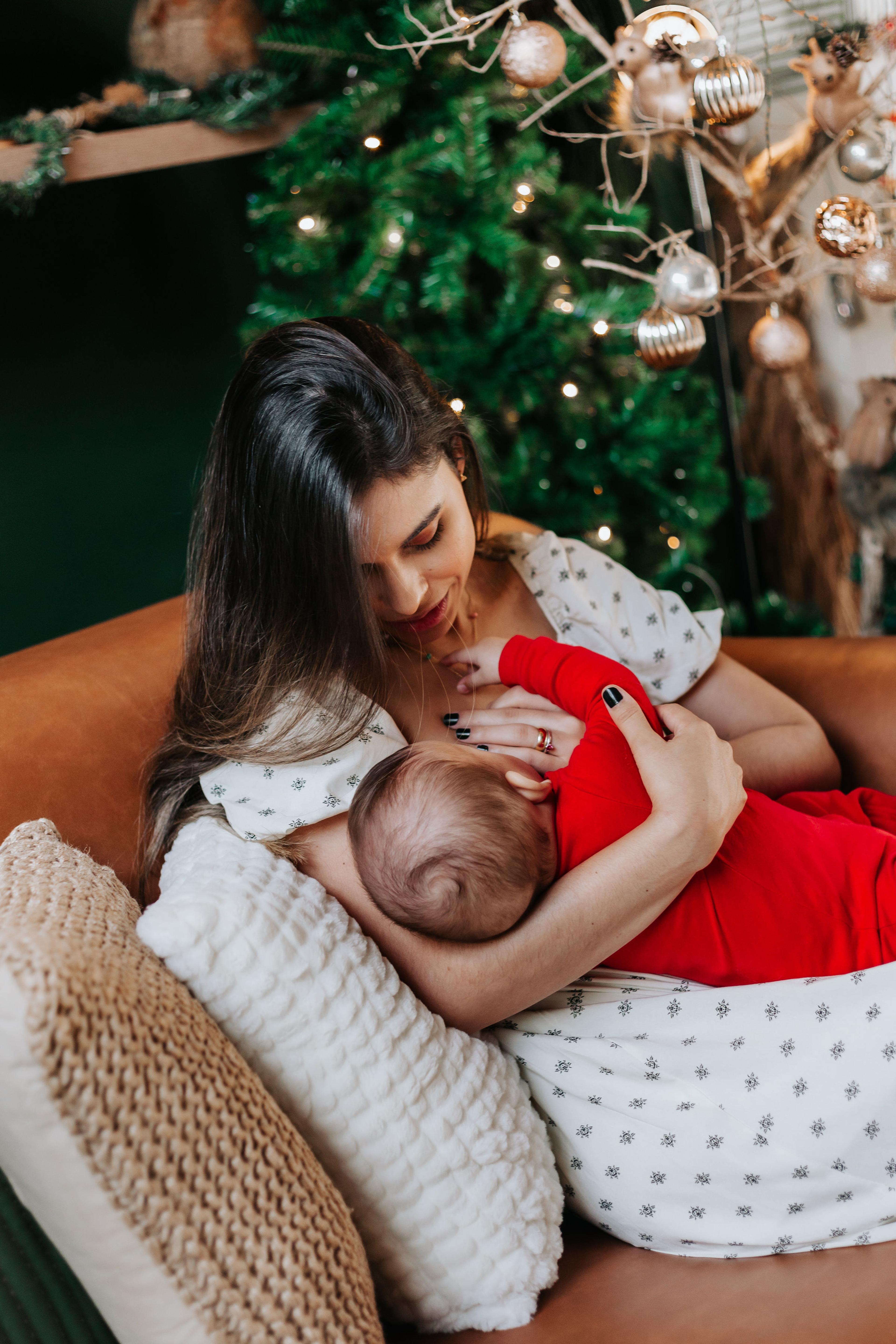 Mãe amamenta o bebê de body vermelho ao lado da árvore de Natal em primeiro plano