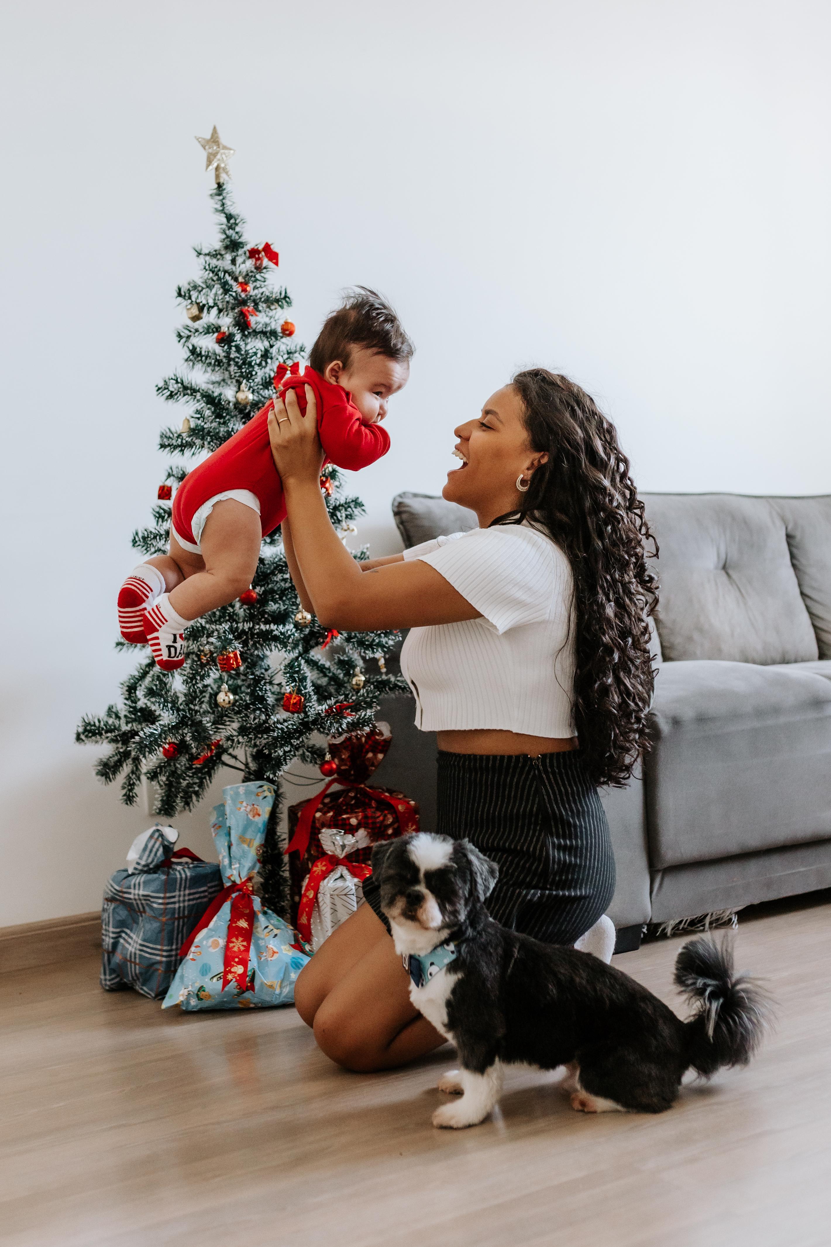 Mãe ergue o bebê de body vermelho ao lado da árvore de Natal, com o cachorro da família em primeiro plano