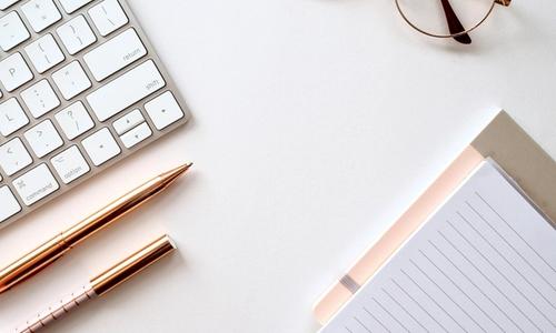 imagine facuta de sus asupra unei mese pe care e o tastatura, doua pixuri, ochelari si un blocknote
