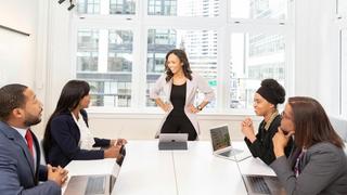 Meeting of four persons with laptops at a table and one standing and smiling