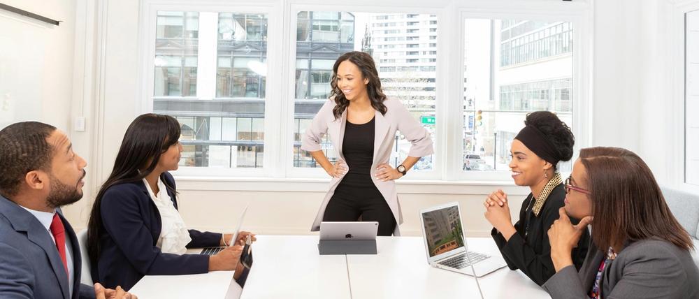 Meeting of four persons with laptops at a table and one standing and smiling