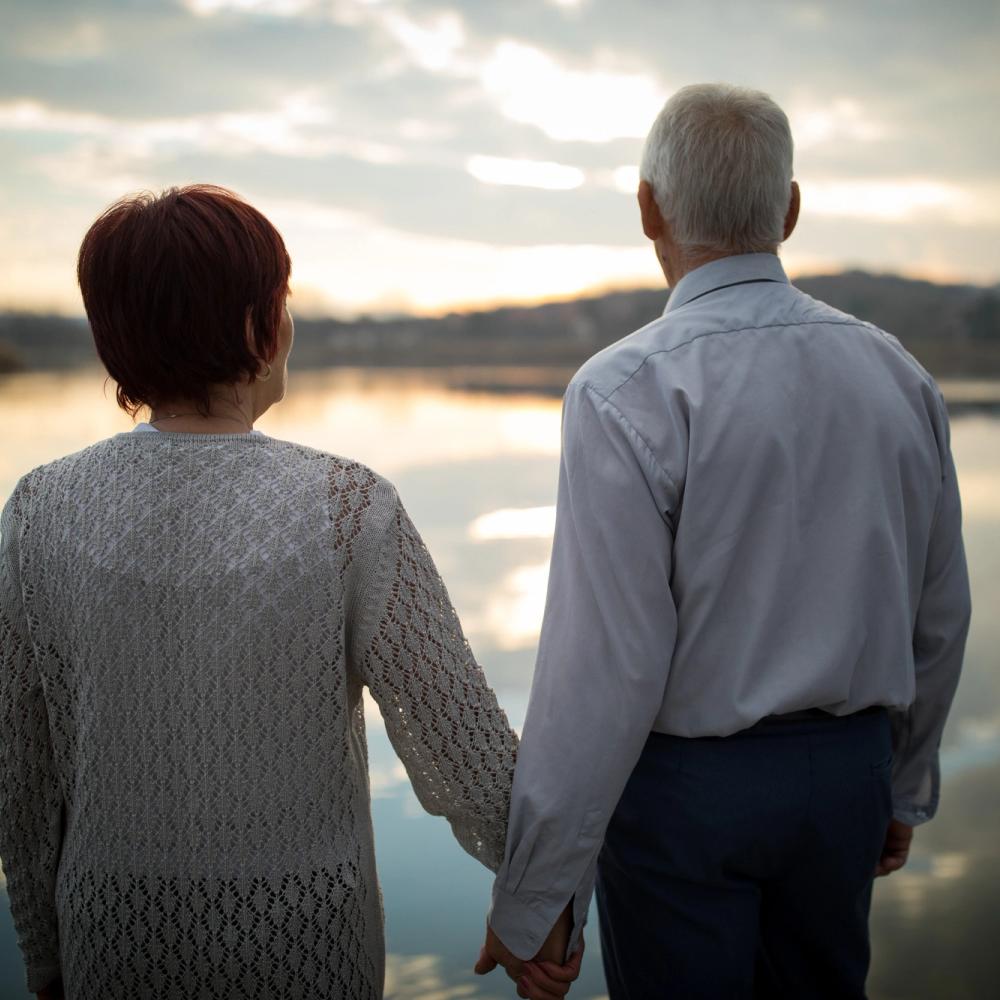 To eldre som holder hender og ser utover et vann. Foto.