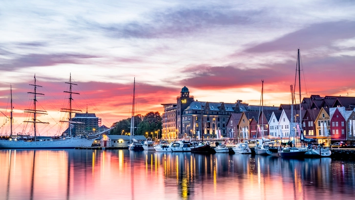 Bergen's historic Bryggen wharf with the Statsraad Lehmkuhl at sunset Sunset view of Bergen's historic Bryggen wharf with colorful wooden buildings and the tall ship Statsraad Lehmkuhl moored in the harbor, with dramatic pink and purple sky reflecting in calm waters