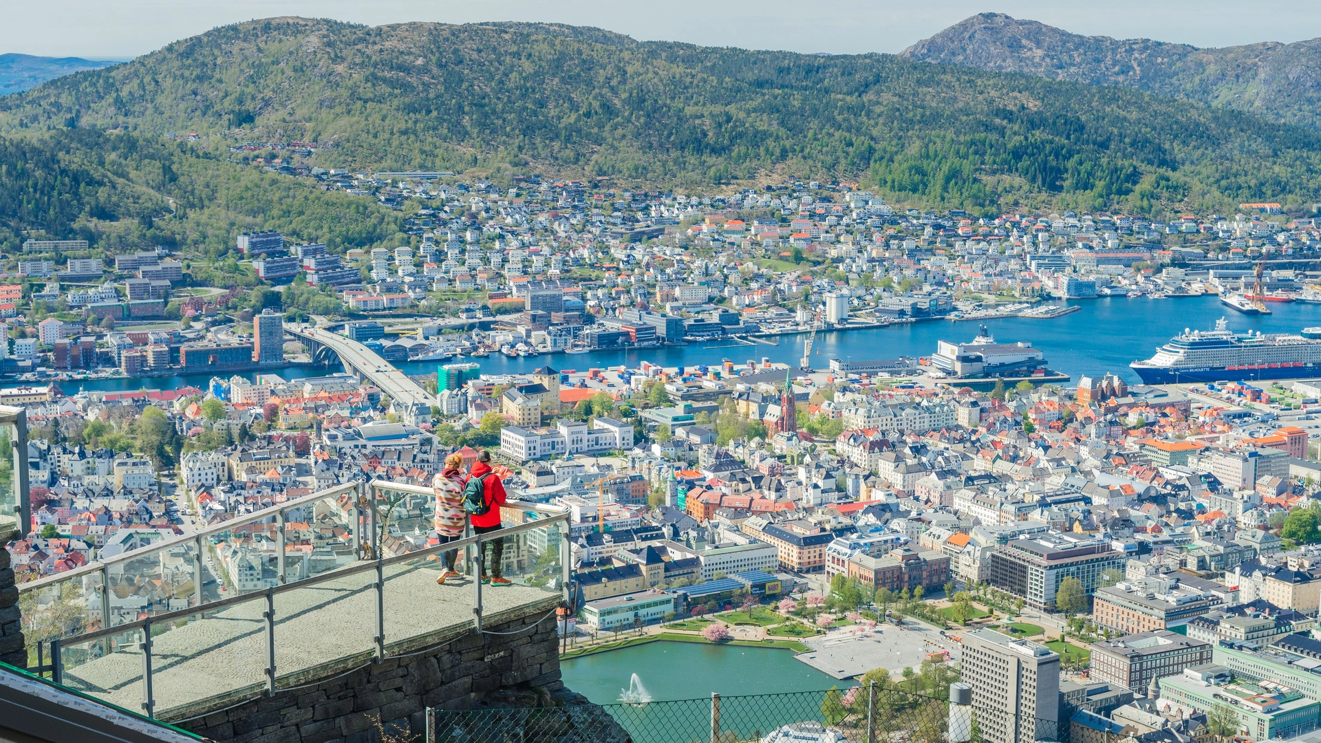 Spring view from Mount Fløyen over Bergen Panoramic view from Mount Fløyen showing Bergen city center, harbor, and surrounding mountains in spring, with a person in red jacket on the viewing platform overlooking the scenic landscape.
