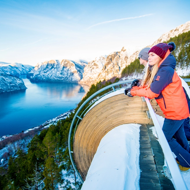Frau am Aussichtspunkt Stegastein über dem Aurlandsfjord im Winter Frau genießt spektakuläre Panoramaaussichten von der schneebedeckten Aussichtsplattform Stegastein über den Aurlandsfjord und dramatische norwegische Berge - als Zusatzoption zur Norway in a Nutshell Wintertour verfügbar