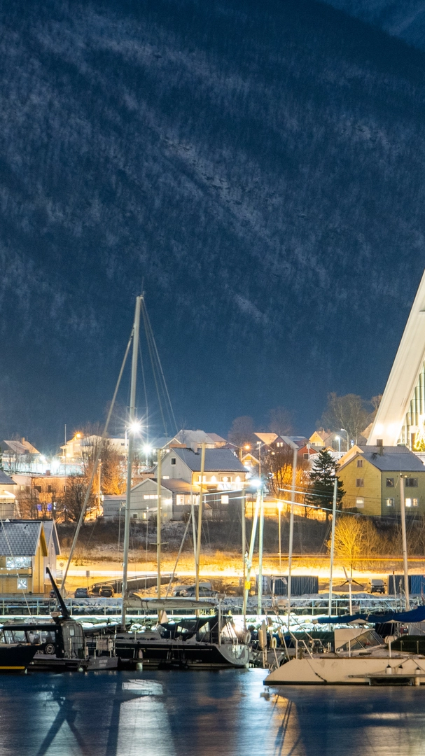 Ishavskatedralen i kveldslys Den opplyste Ishavskatedralen i Tromsø om kvelden, med fiskebåter i havnen nedenfor og snødekte fjell som reiser seg i bakgrunnen.
