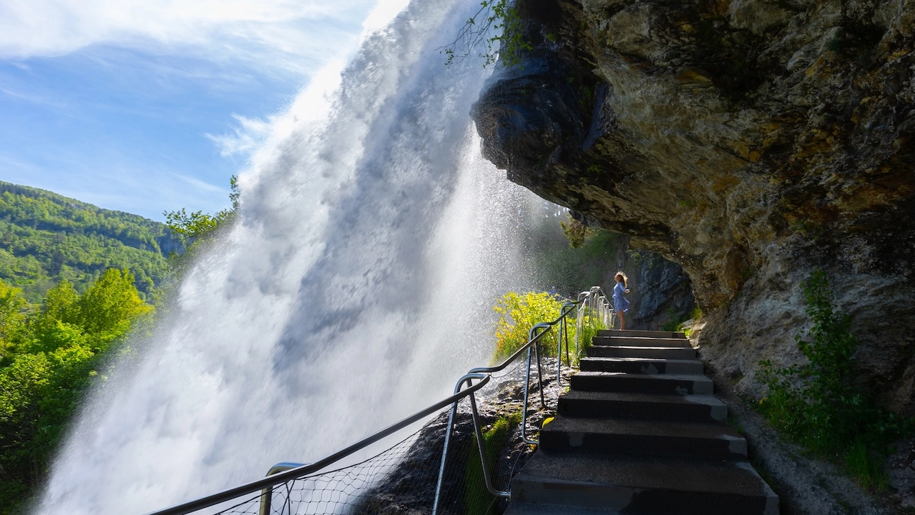 Experience Steinsdalsfossen - Norway's Accessible Spring Waterfall Traveler standing behind the powerful Steinsdalsfossen waterfall surrounded by lush green spring vegetation and dramatic rocky cliffs in Norway