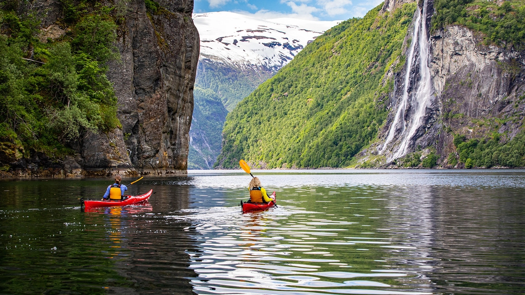 Kayaking beneath the Seven Sisters waterfall in Geirangerfjord Two kayakers paddle in red kayaks on calm fjord waters with the dramatic Seven Sisters waterfall cascading down steep green mountainsides and snow-capped peaks in the background