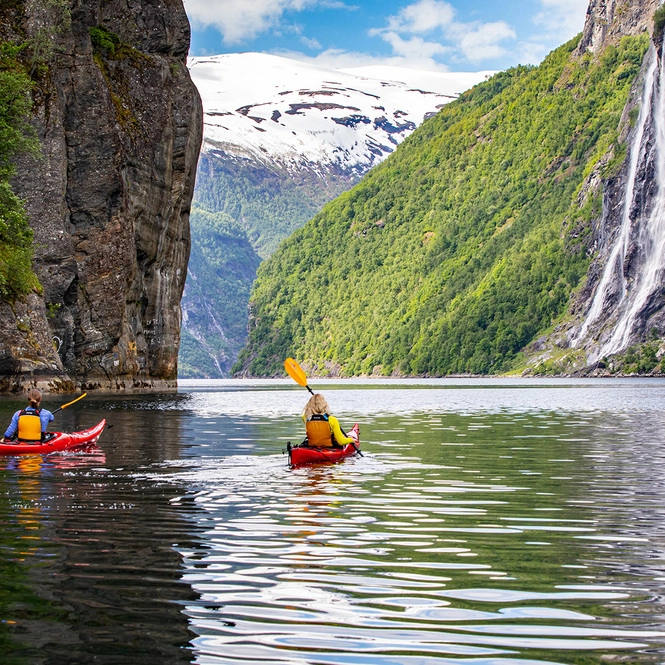 Kajakk under De syv søstre-fossen i Geirangerfjorden To kajakpadlere i røde kajakker på rolige fjordvann med det dramatiske De syv søstre-fossen som styrter ned bratte grønne fjellsider med snødekte topper i bakgrunnen
