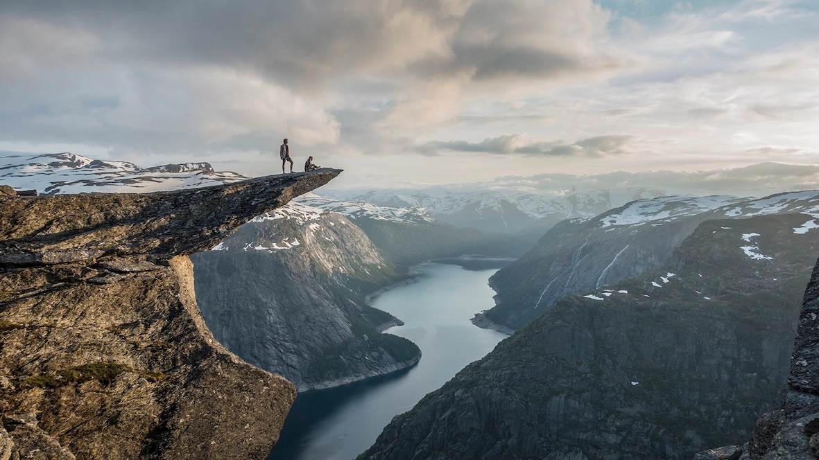 Trolltunga hiking adventure with snow-capped mountain panorama in Norway Hikers standing on Trolltunga rock formation overlooking Ringedalsvatnet lake surrounded by snow-capped mountains in Norway