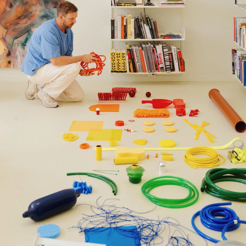 A designer crouches on a studio floor arranging blue, green, and yellow hoses alongside wires, filters, and industrial components