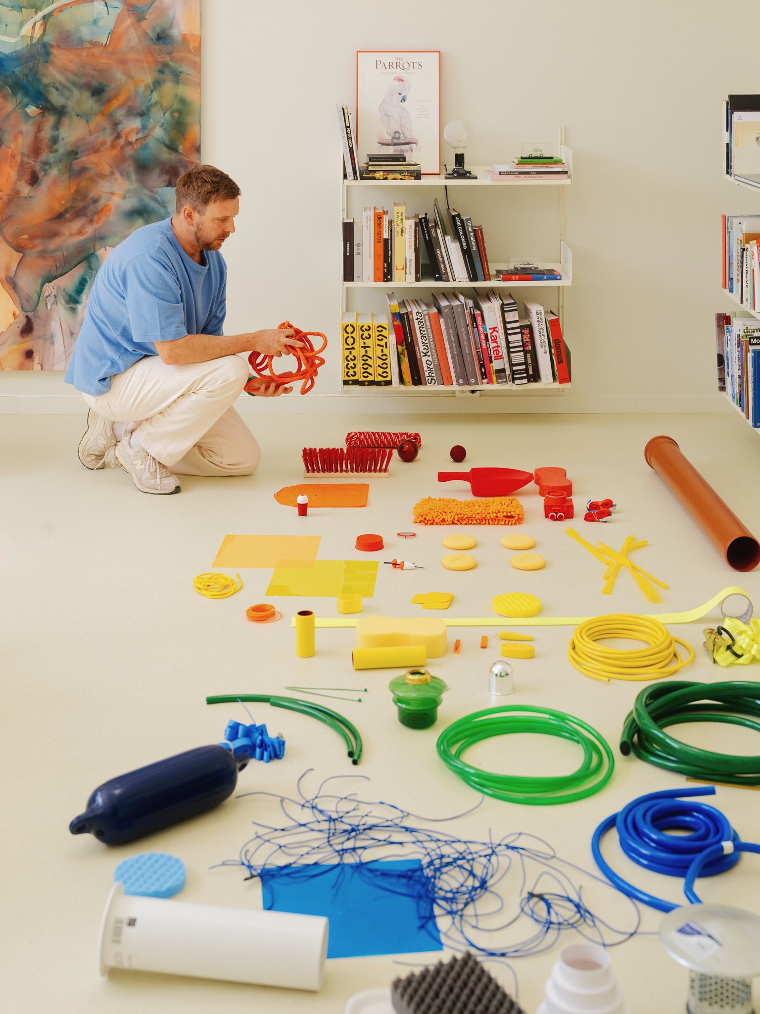 A designer crouches on a studio floor arranging blue, green, and yellow hoses alongside wires, filters, and industrial components