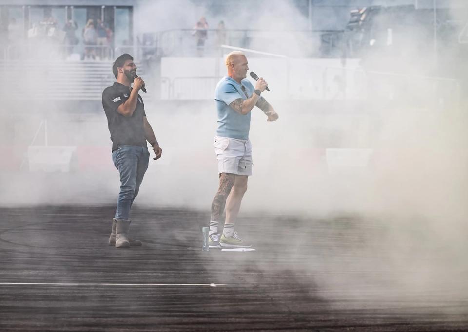 Two presenters with microphones standing in a cloud of tyre smoke