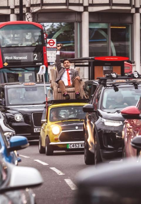 Sam dressed as Mr Bean, riding an armchair on the roof of a yellow Mini through London traffic