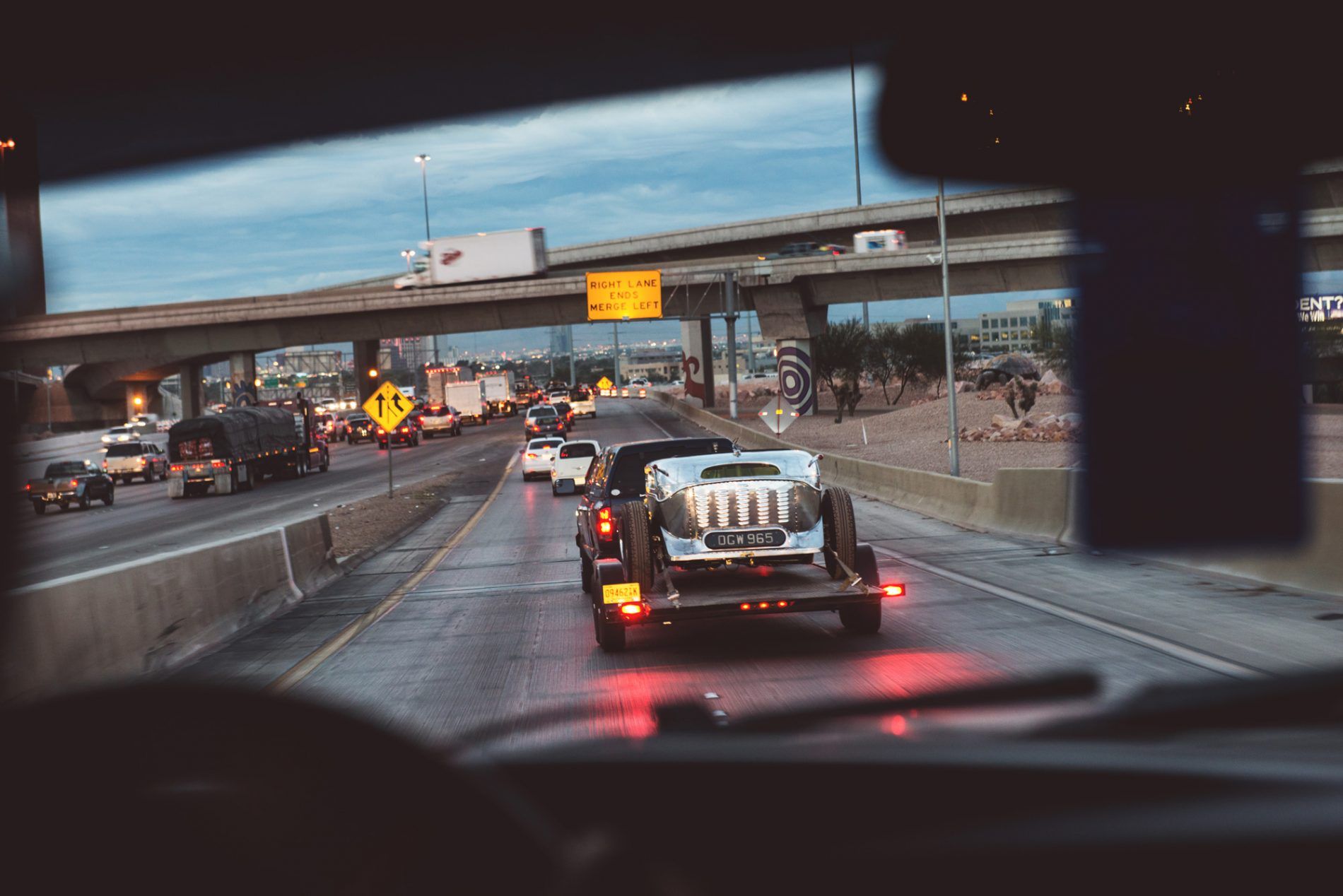 A hot rod on a trailer, seen through the windscreen on a motorway at dusk