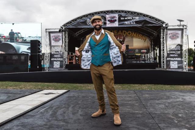 Sam on stage at a car show in a waistcoat and flat cap, arms spread wide
