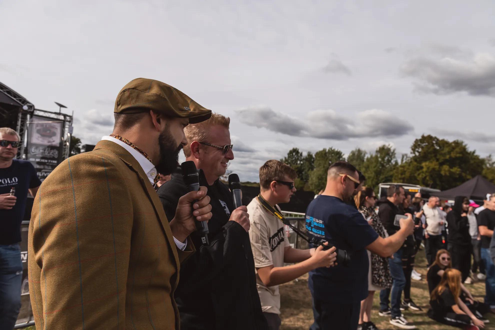 Sam in a tweed suit and flat cap talking to a crowd at a car show