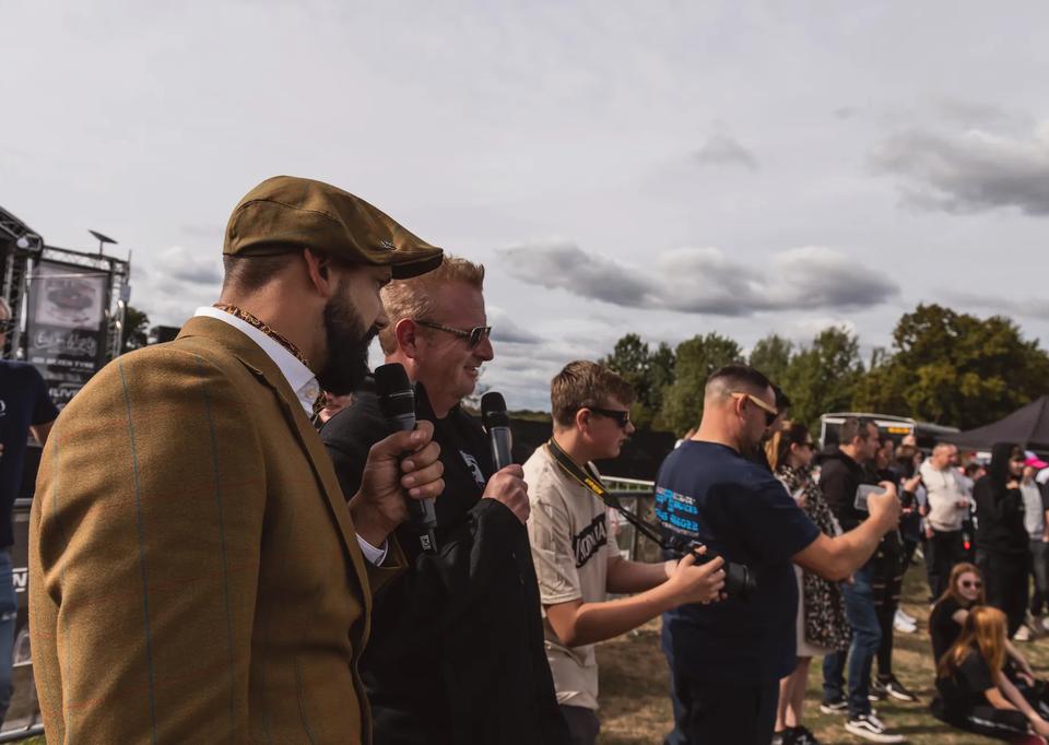 Sam in a tweed suit and flat cap talking to a crowd at a car show