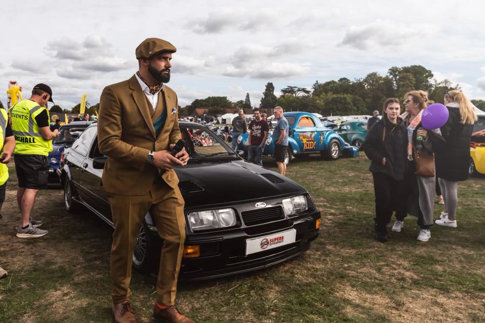 Sam in a tweed suit leaning on a black Ford Sierra at a car show