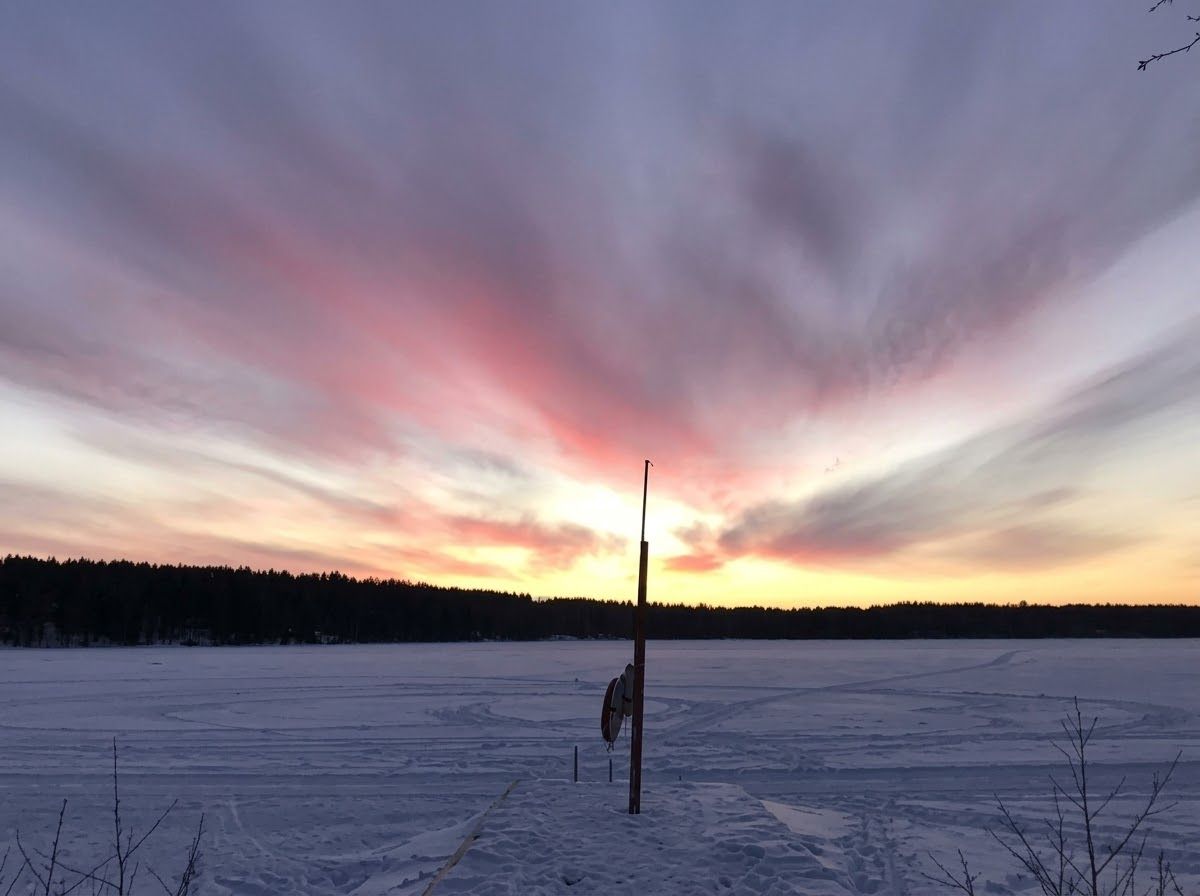 Et vidstrakt landskapsfotografi av en slående vintersolnedgang. Himmelen dominerer den øvre halvdelen med dramatiske striper av rosa, magenta og gule skyer. Nedenfor strekker en mørk skogslinje seg over horisonten. Forgrunnen viser en frossen innsjø helt dekket av snø, som viser ski- og fotspor i flere retninger. En høy, tynn trestang står fremtredende på dekket i forgrunnen nederst til høyre, som livbøyen som er festet til den, og nakne grener stikker opp av snøen i de nederste hjørnene. A wide landscape photograph of a striking winter sunset. The sky dominates the upper half with dramatic streaks of pink, magenta, and yellow clouds. Below, a dark forest line stretches across the horizon. The foreground features a frozen lake entirely covered in snow, showing ski and foot tracks in several directions. A tall, thin wooden pole stands prominently of the deck in the lower right foreground, as the life-buoy attached to it, and bare branches poke out of the snow in the bottom corners.