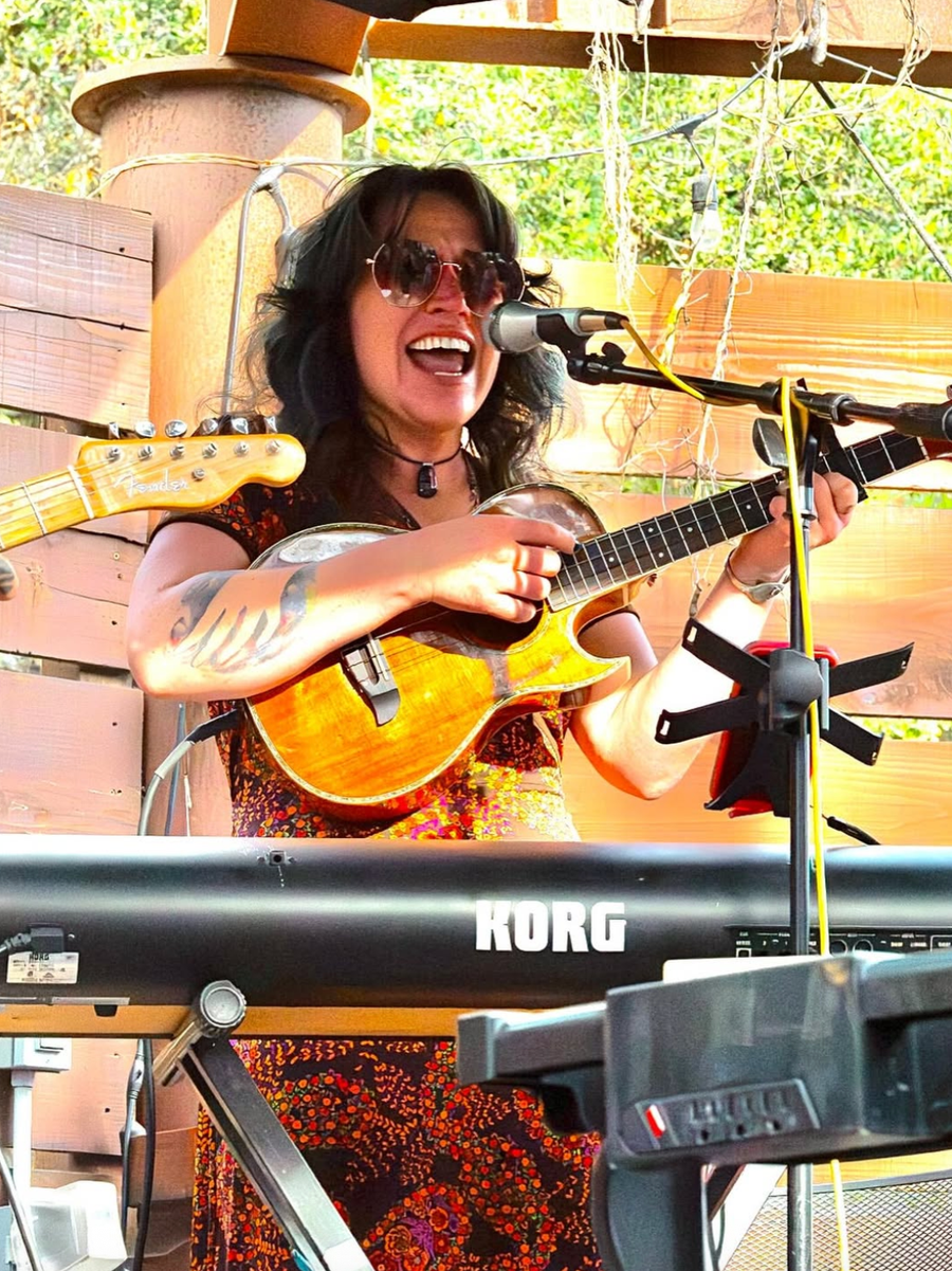 Rachel singing into a microphone while playing a baritone ukulele at an outdoor show.