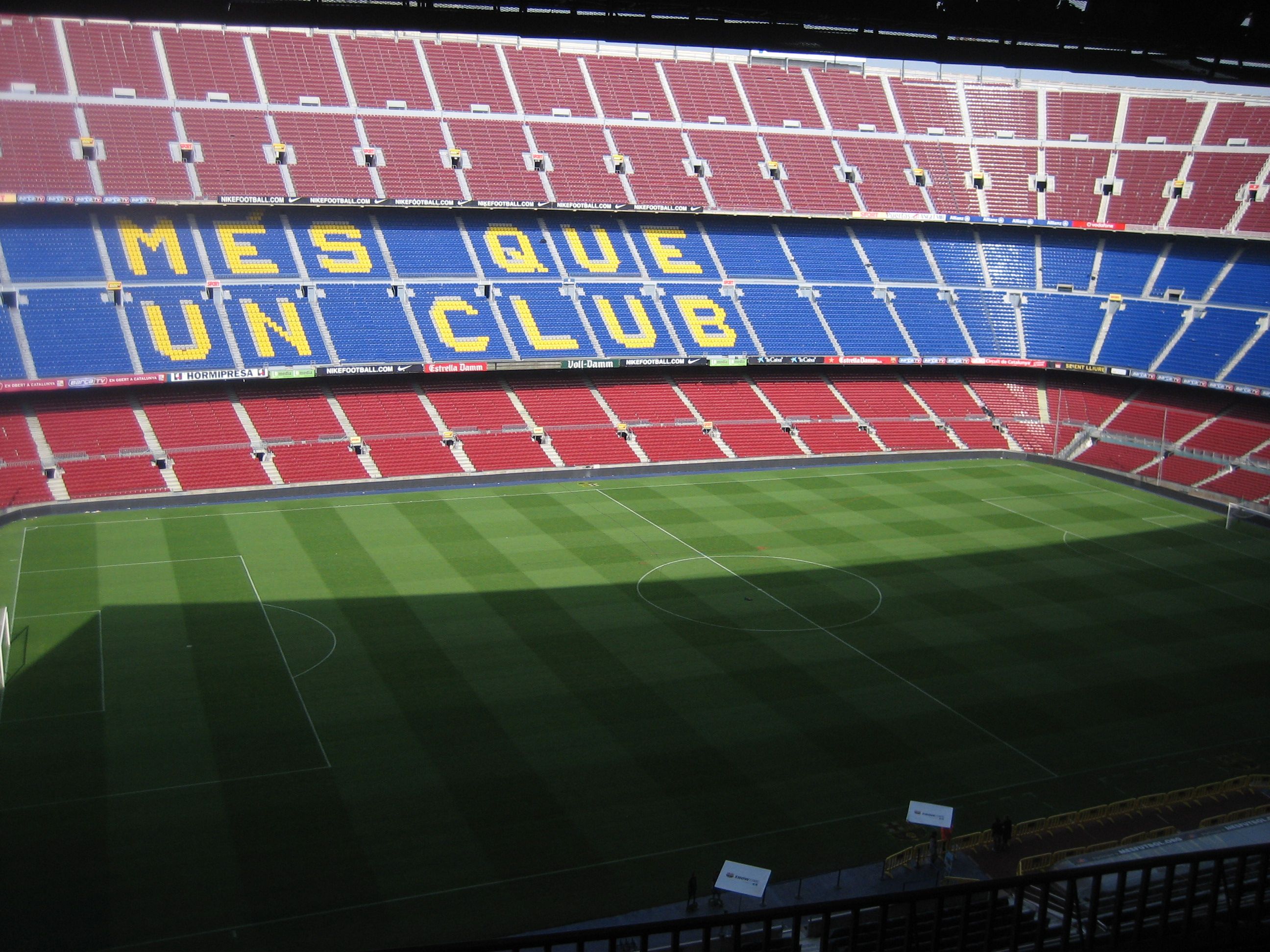 Estadio de fútbol con asientos rojos y azules, campo de hierba verde y lema visible en la zona de gradas