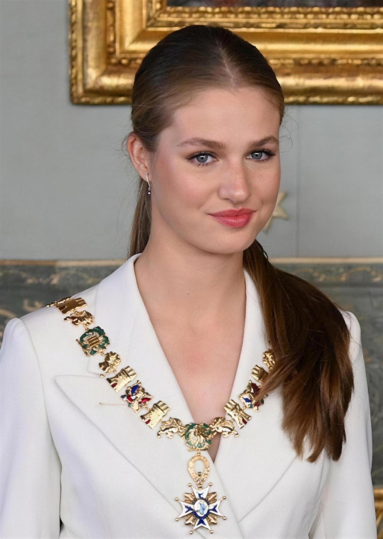 Mujer de perfil con traje blanco y collar ceremonial dorado con medallas, ante marco dorado en pared clara