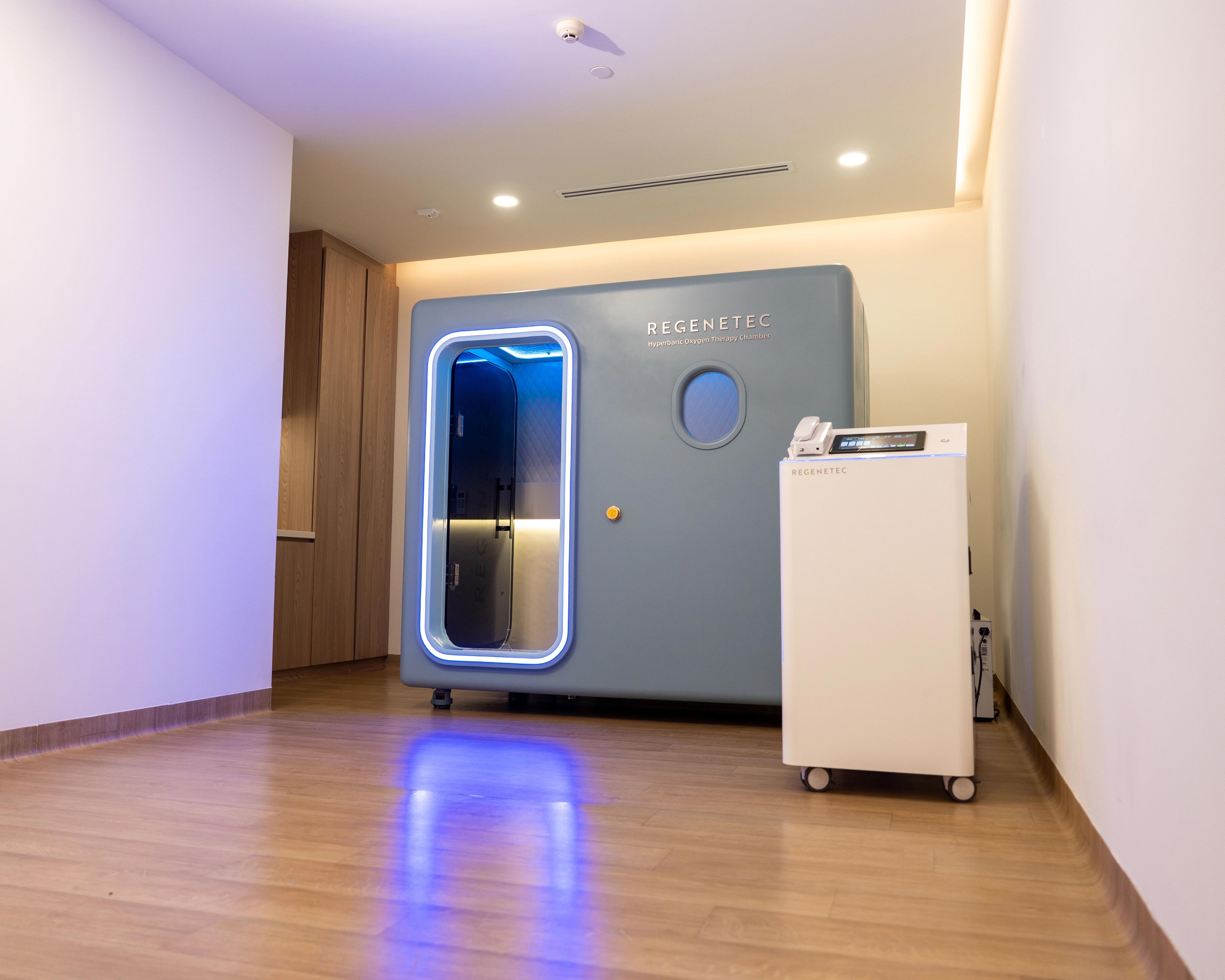Blue HBOT machine with lights around the door and a small window, inside a room at a medical facility.
