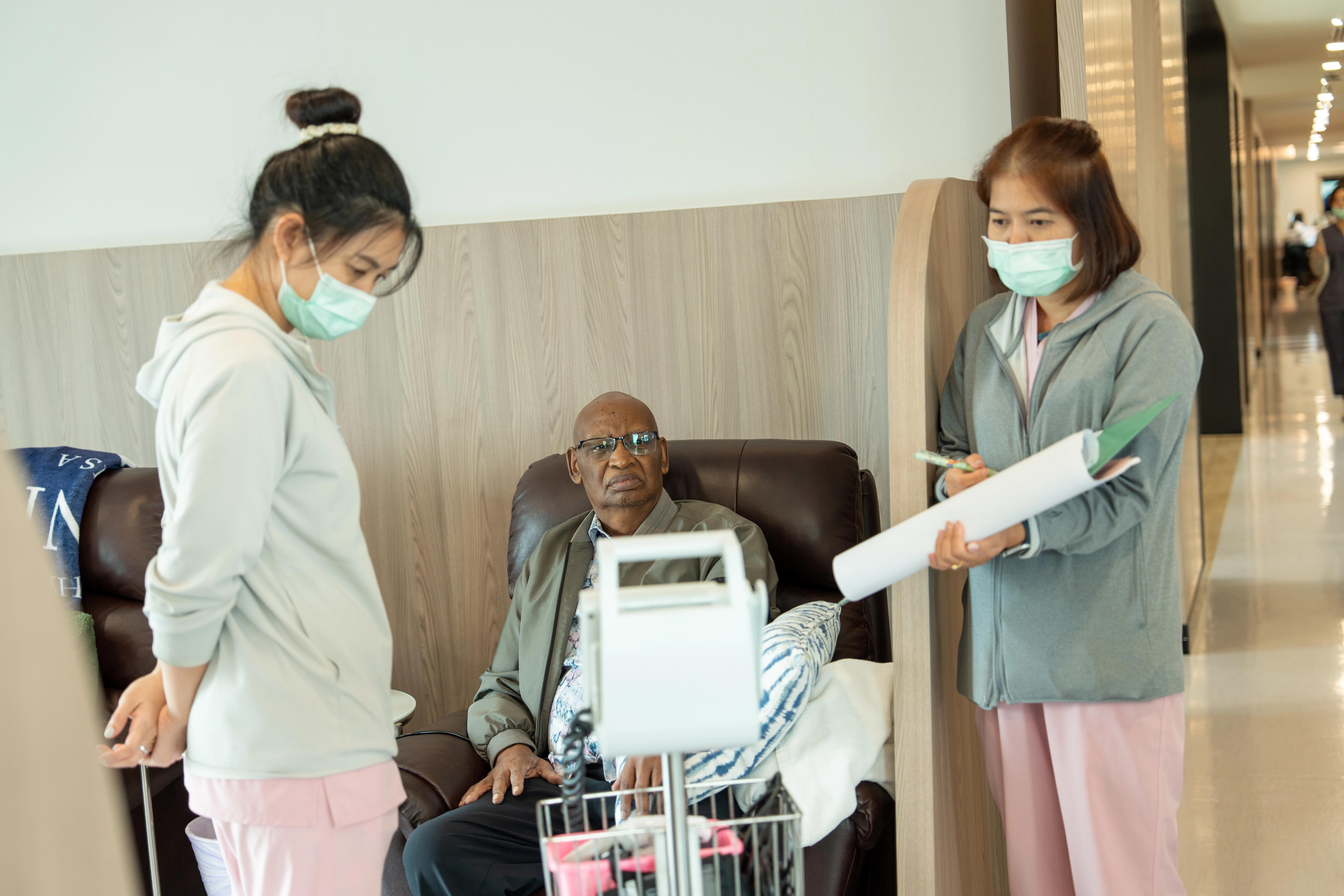 Two nurses stand checking the vitals of a patient who is seated in a black, leather, armchair.