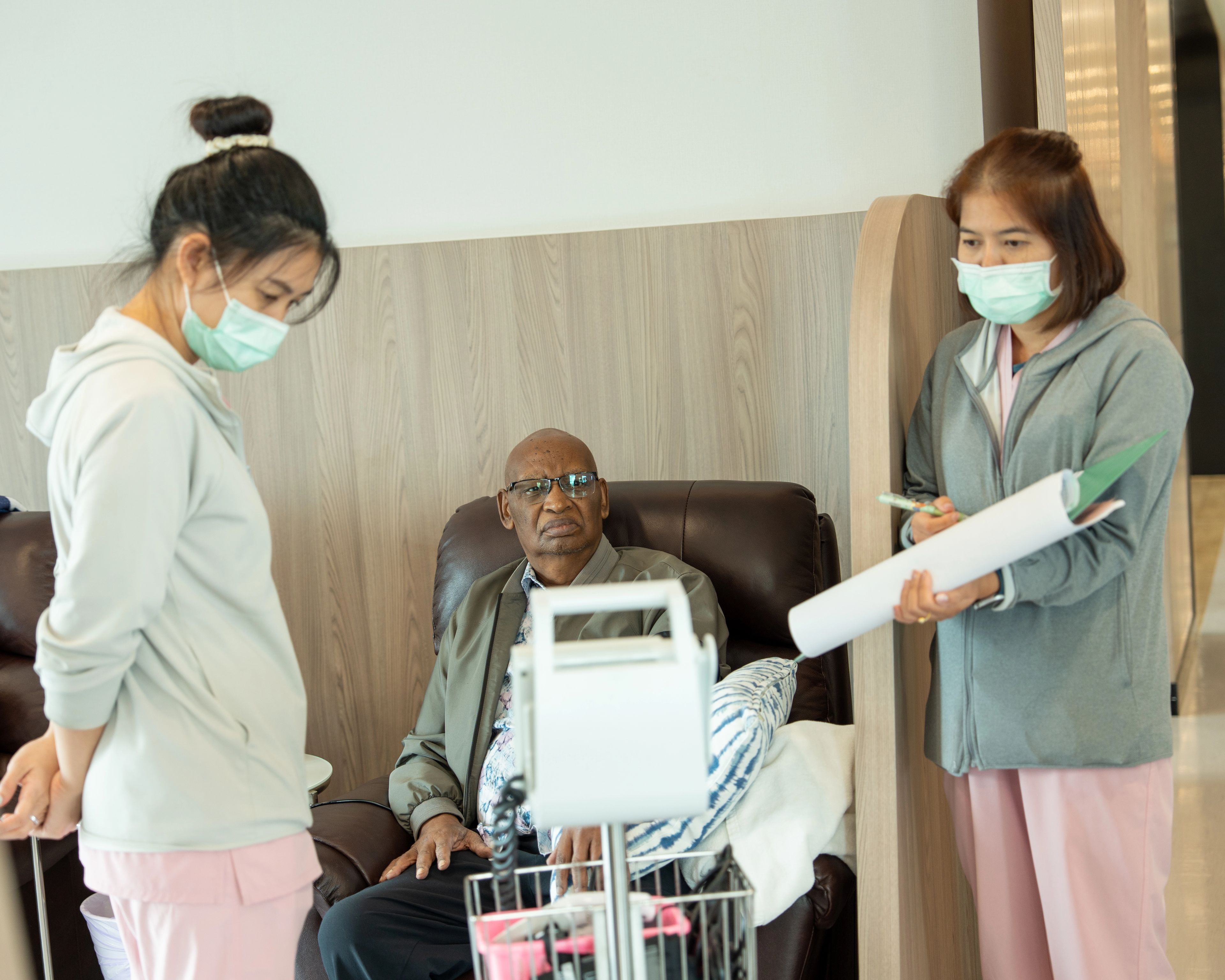 Two nurses stand checking the vitals of a patient who is seated in a black, leather, armchair.