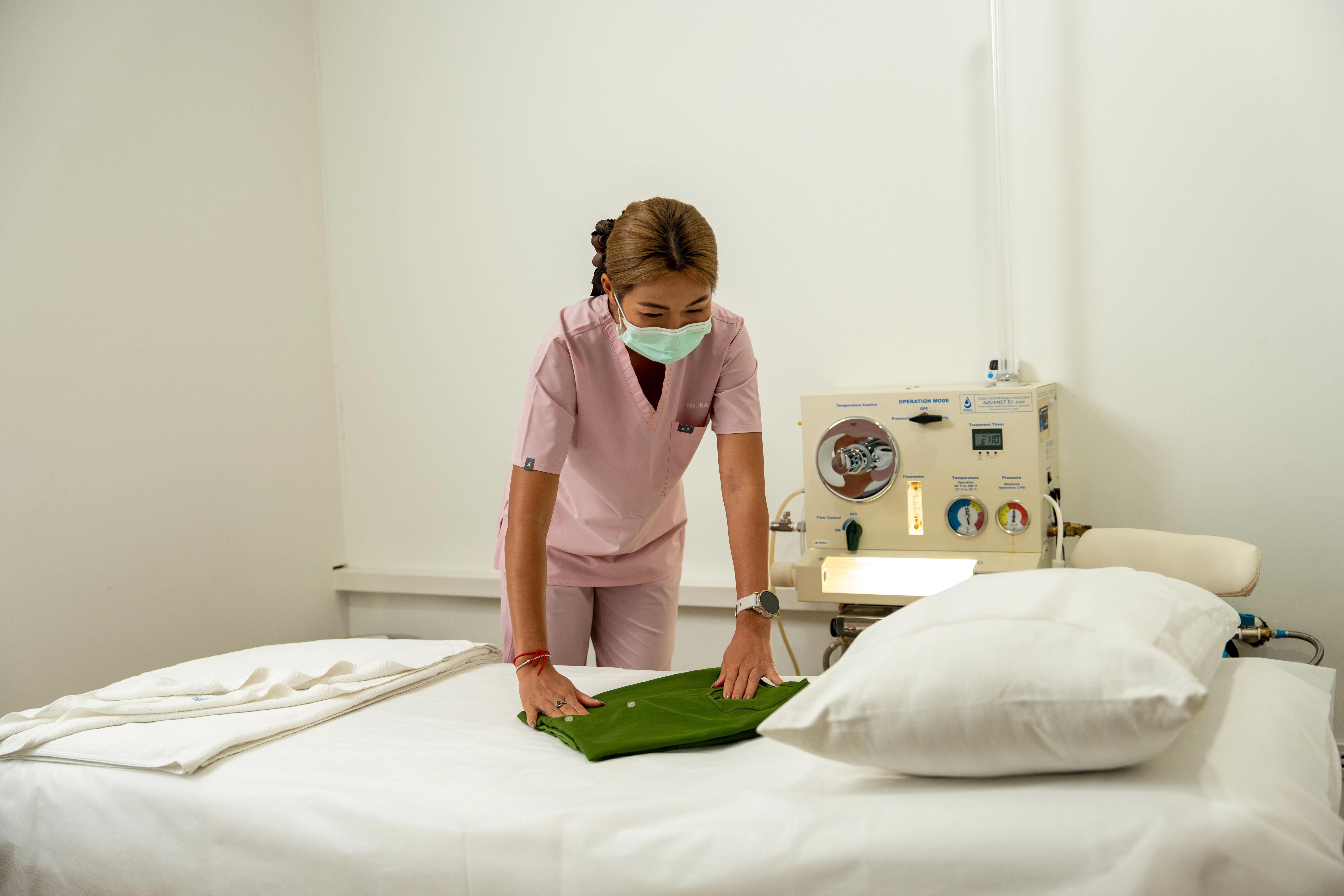 Nurse in pink uniform places green towel on a bed with a lit machine in the background