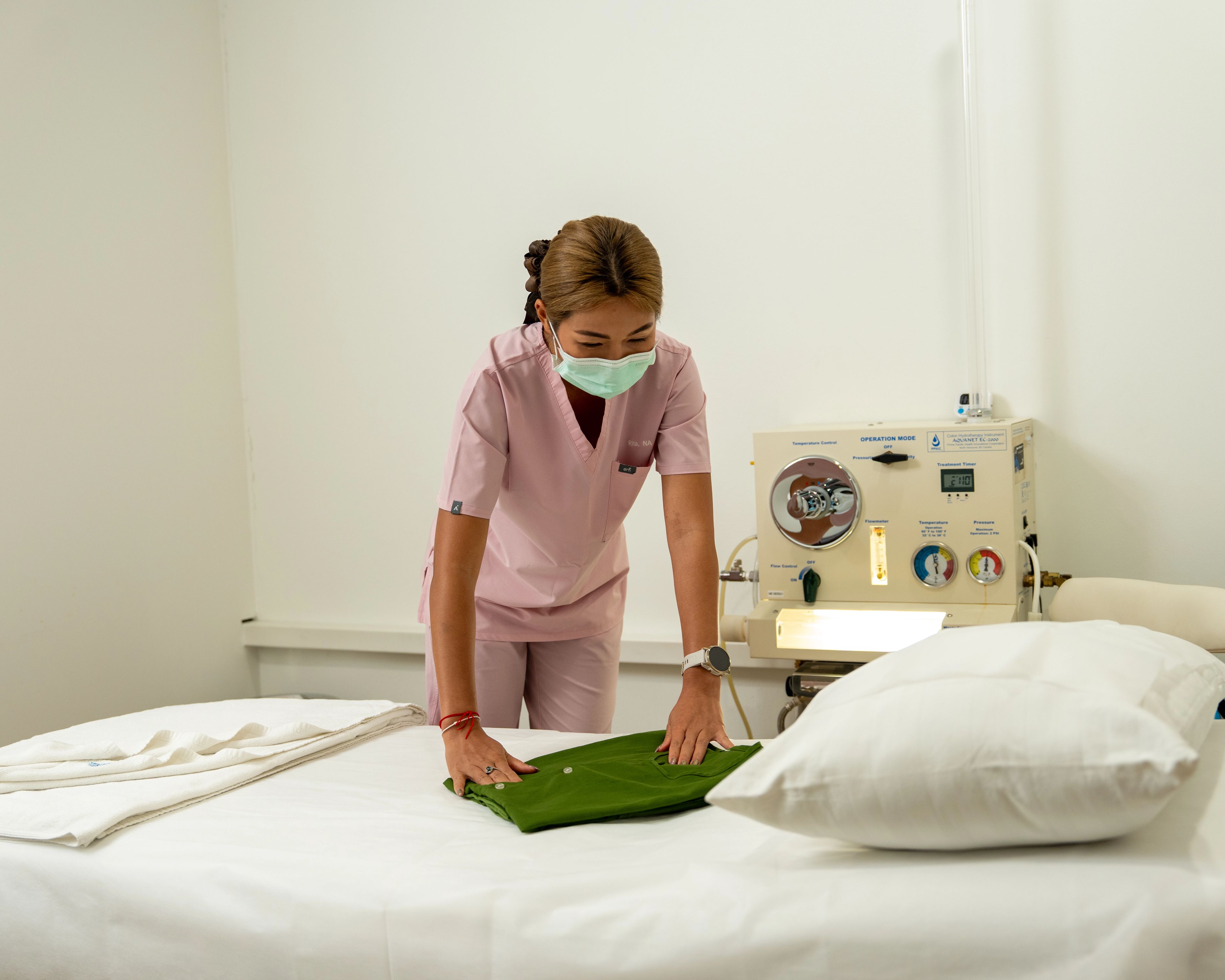 Nurse in pink uniform places green towel on a bed with a lit machine in the background