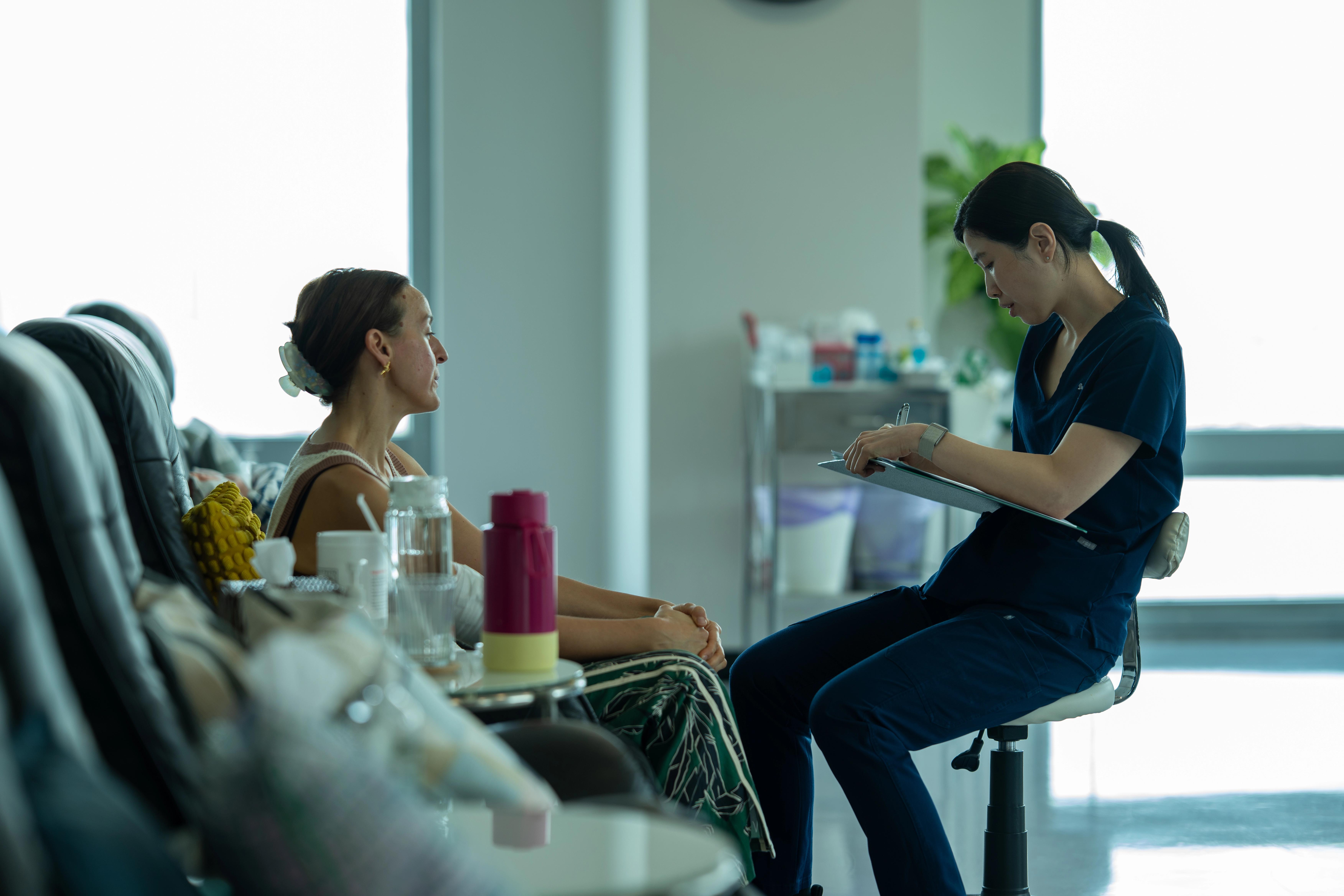 Patient sits in a black leather chair having a conversation with a medical professional in scrubs who is sitting in front of her.