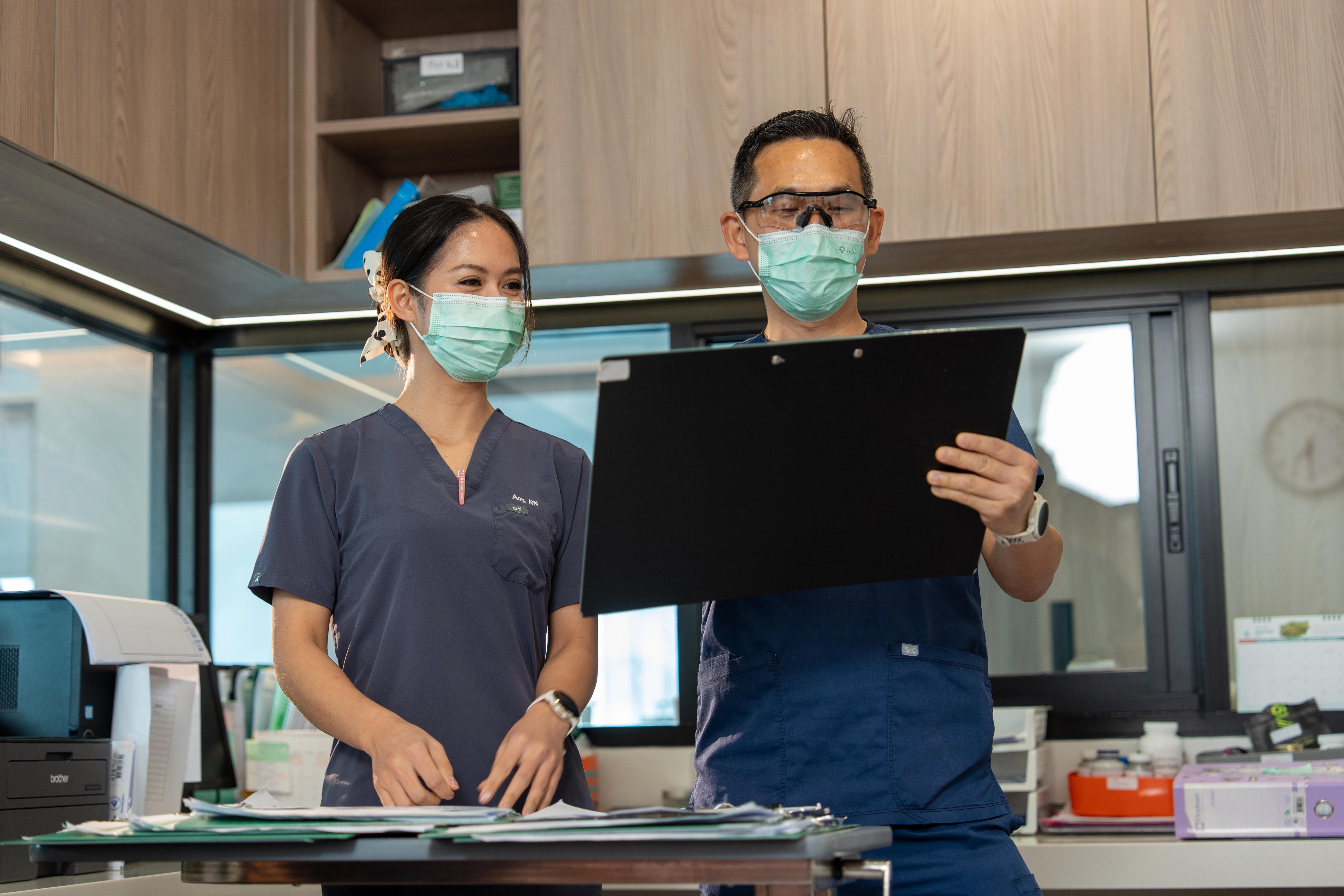 Two medical professionals in scrubs and PPE study a large black clipboard together standing at a desk in an office.