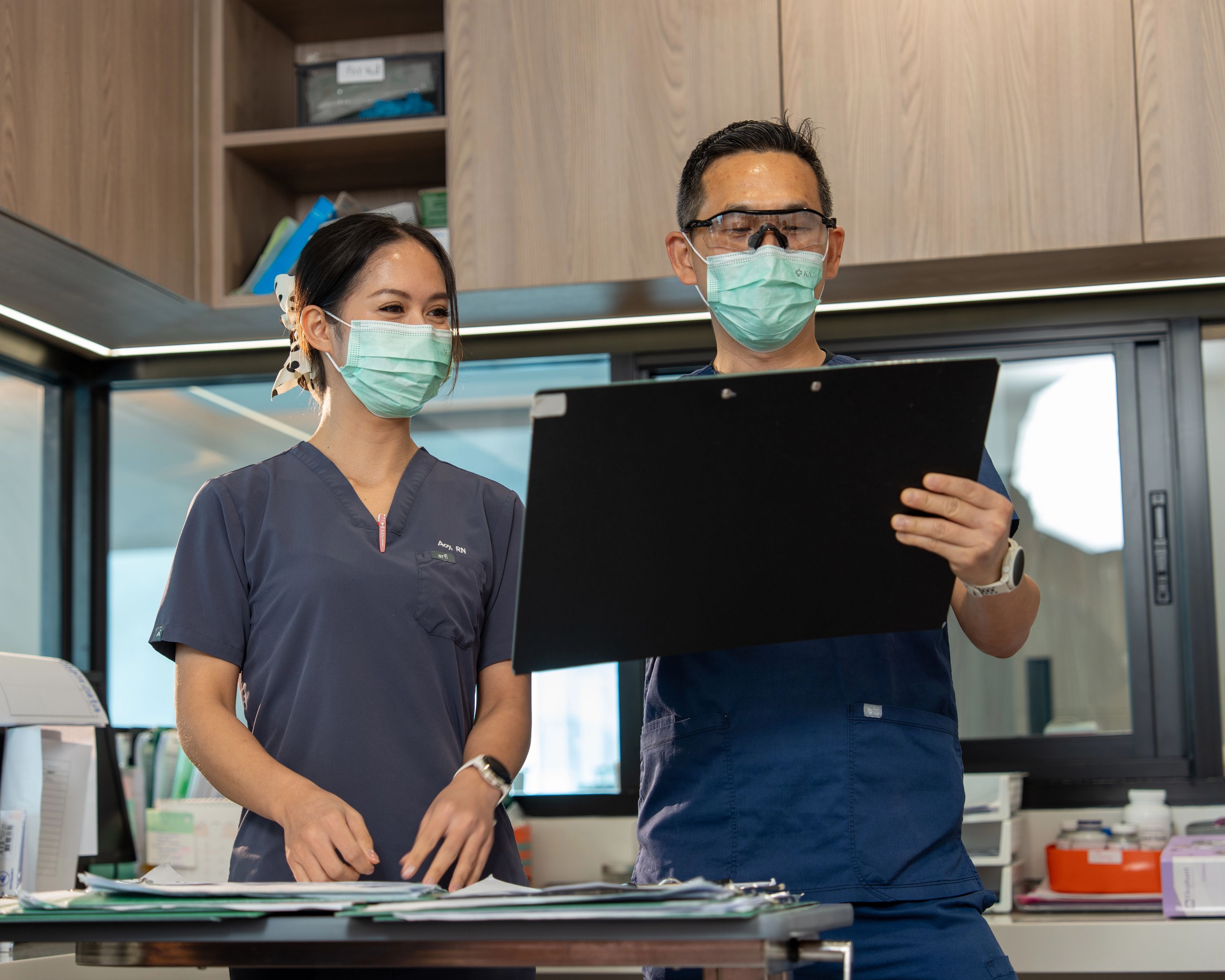 Two medical professionals in scrubs and PPE study a large black clipboard together standing at a desk in an office.