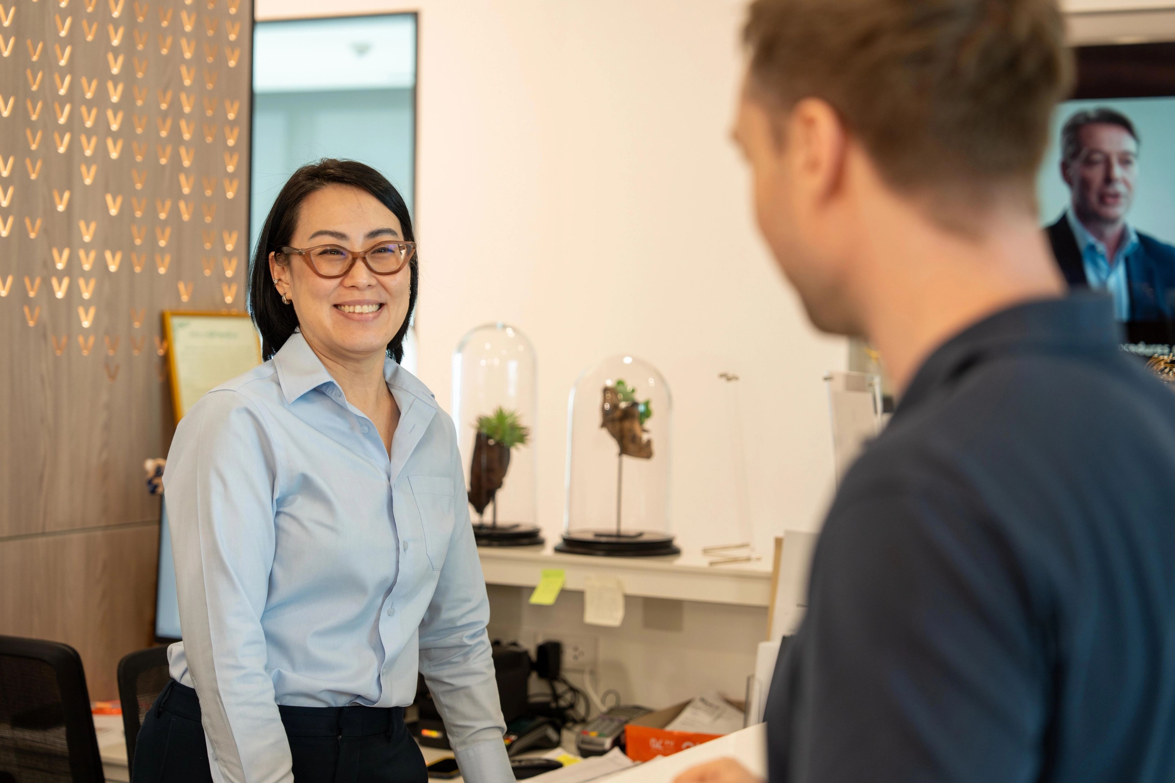 Woman in blue blouse and glasses stands behind a reception desk welcoming a patient to Verita Life.