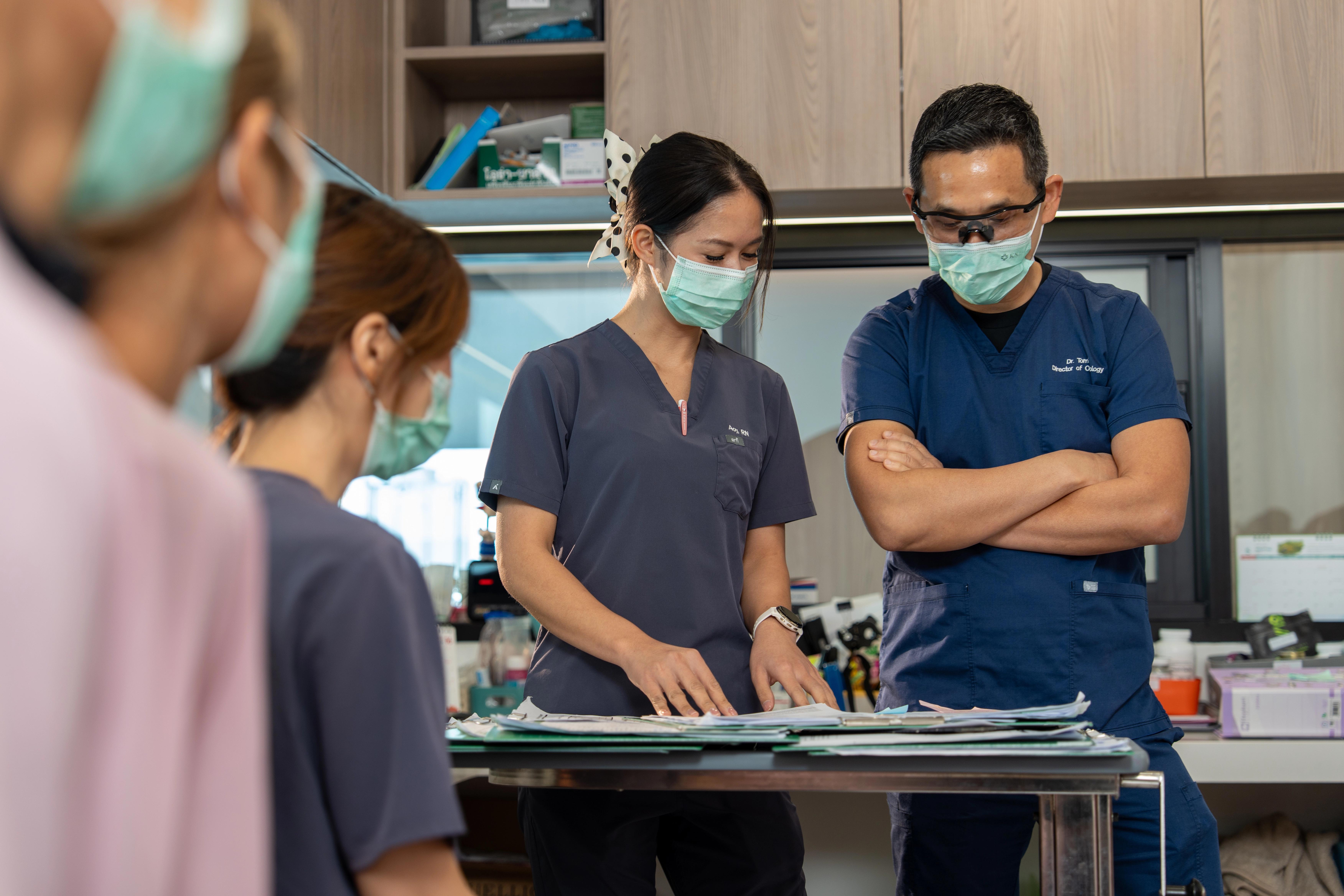 Nurse and doctor in scrubs and face masks having a conversation while looking at notes on a desk in an office, there are three other medical professionals in the room.