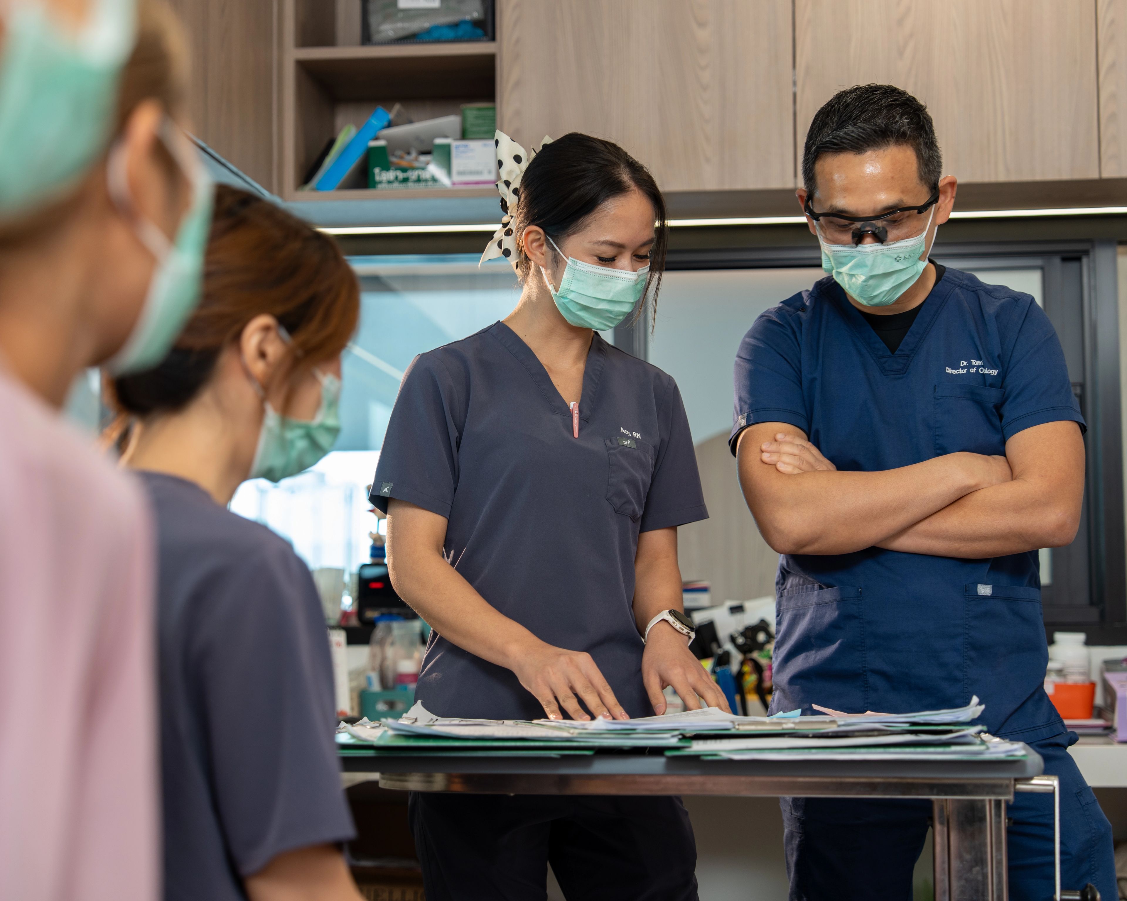 Nurse and doctor in scrubs and face masks having a conversation while looking at notes on a desk in an office, there are three other medical professionals in the room.