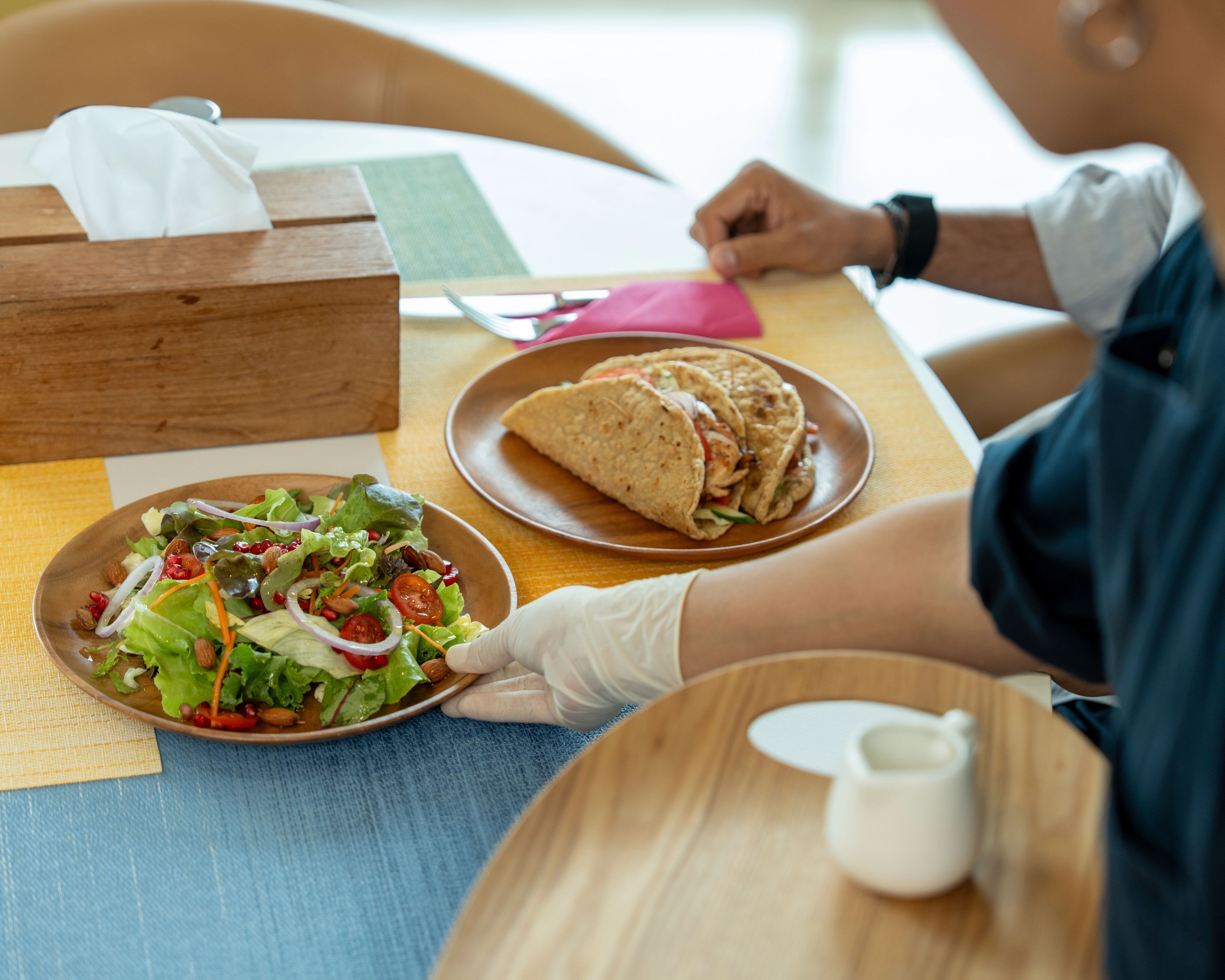A person in white gloves placing a plate of salad on a table next to a plate with two wraps