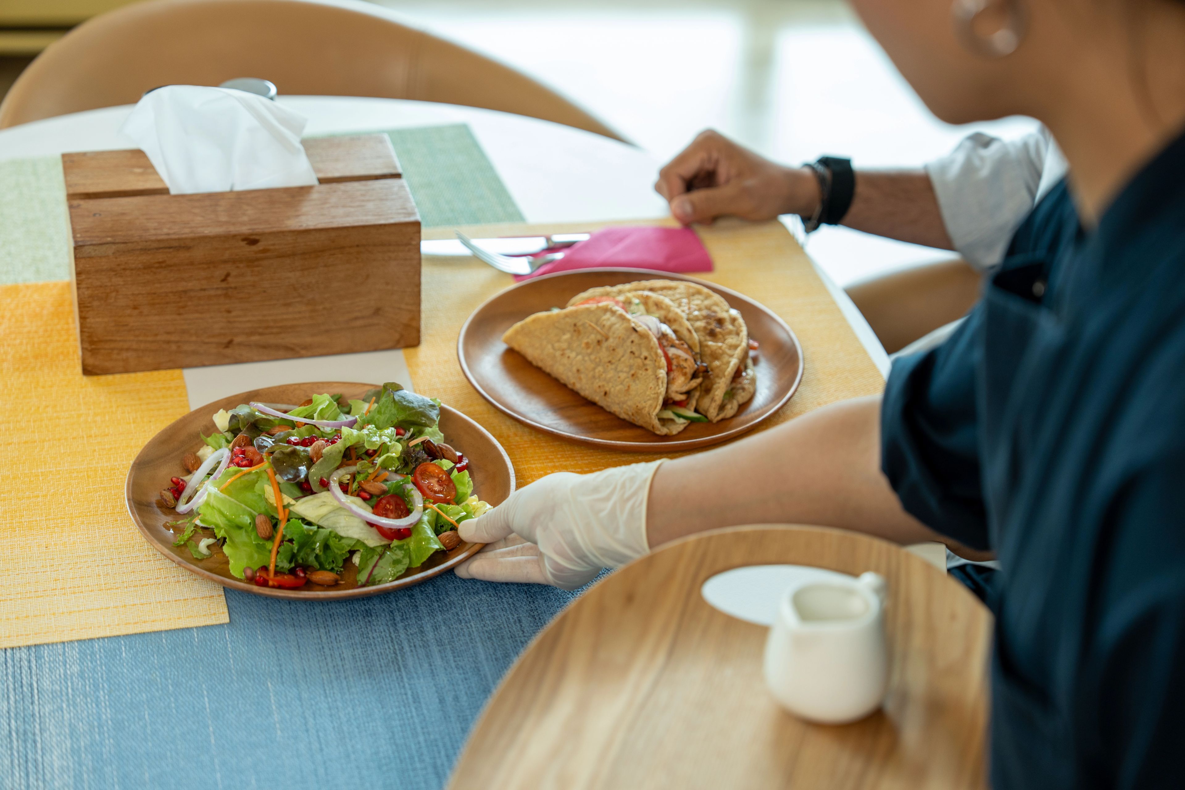 A person in white gloves placing a plate of salad on a table next to a plate with two wraps