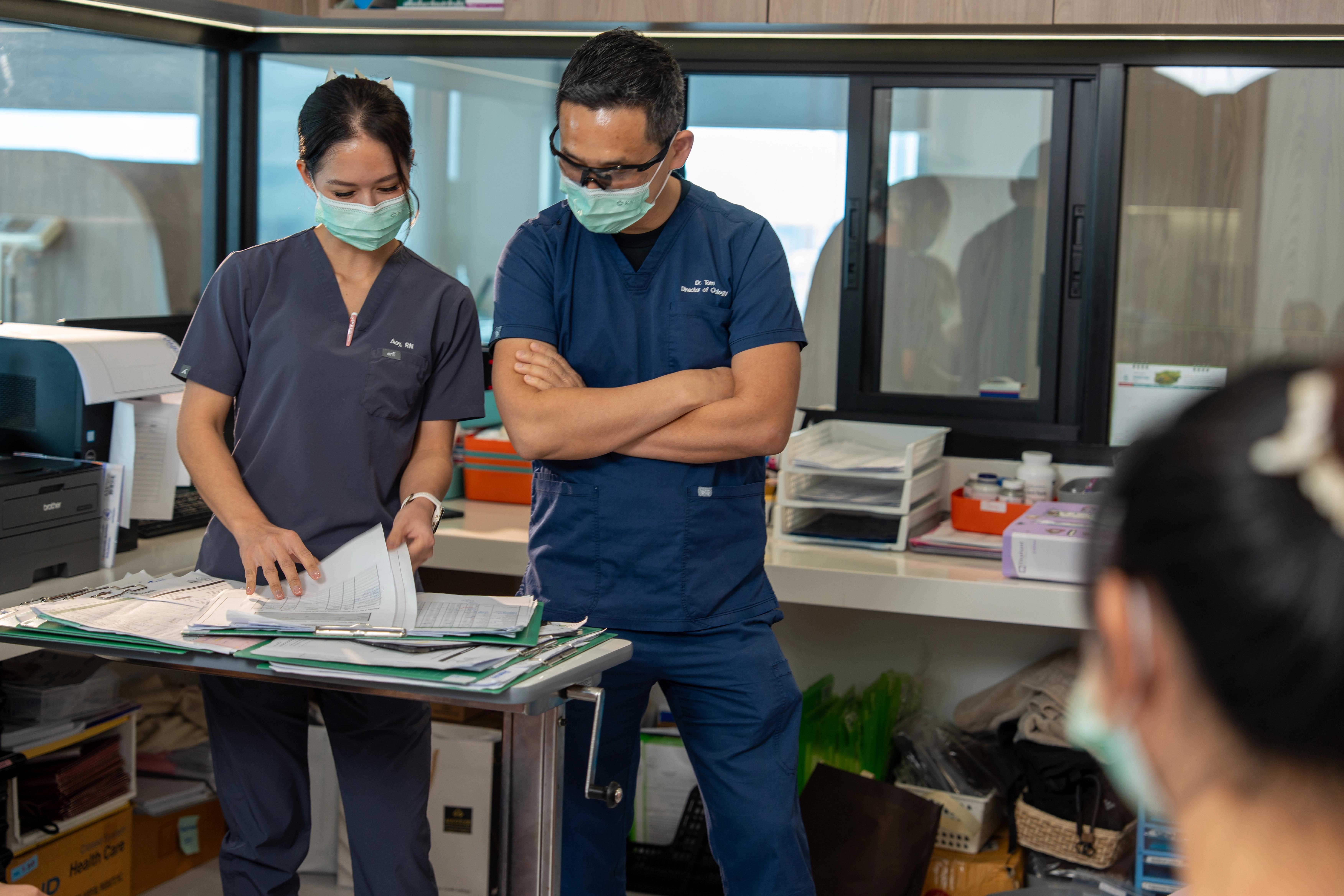 Nurse and doctor in scrubs and face masks having a conversation while looking at notes on a desk in an office, there is a third medical professional in the room.