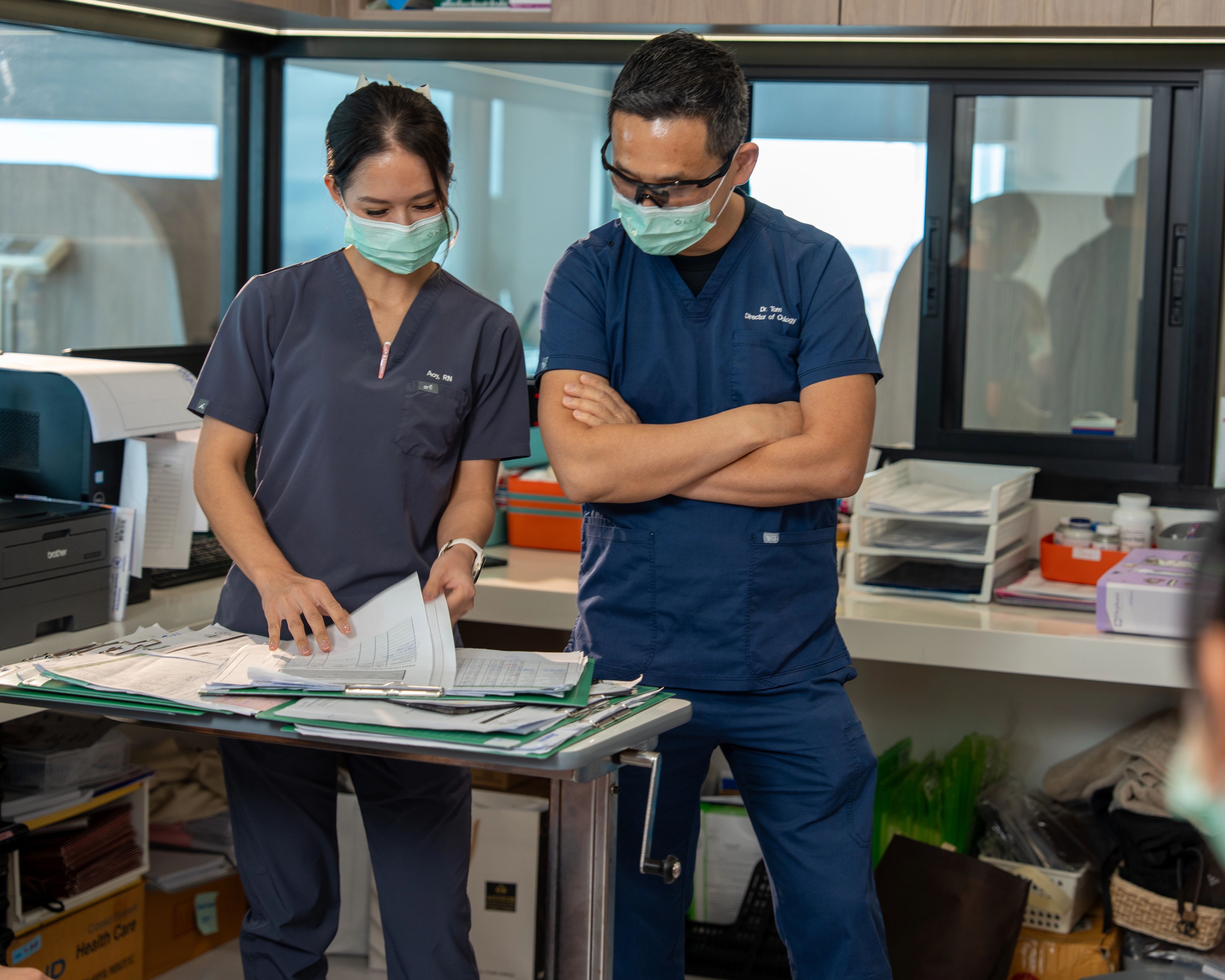 Nurse and doctor in scrubs and face masks having a conversation while looking at notes on a desk in an office, there is a third medical professional in the room.