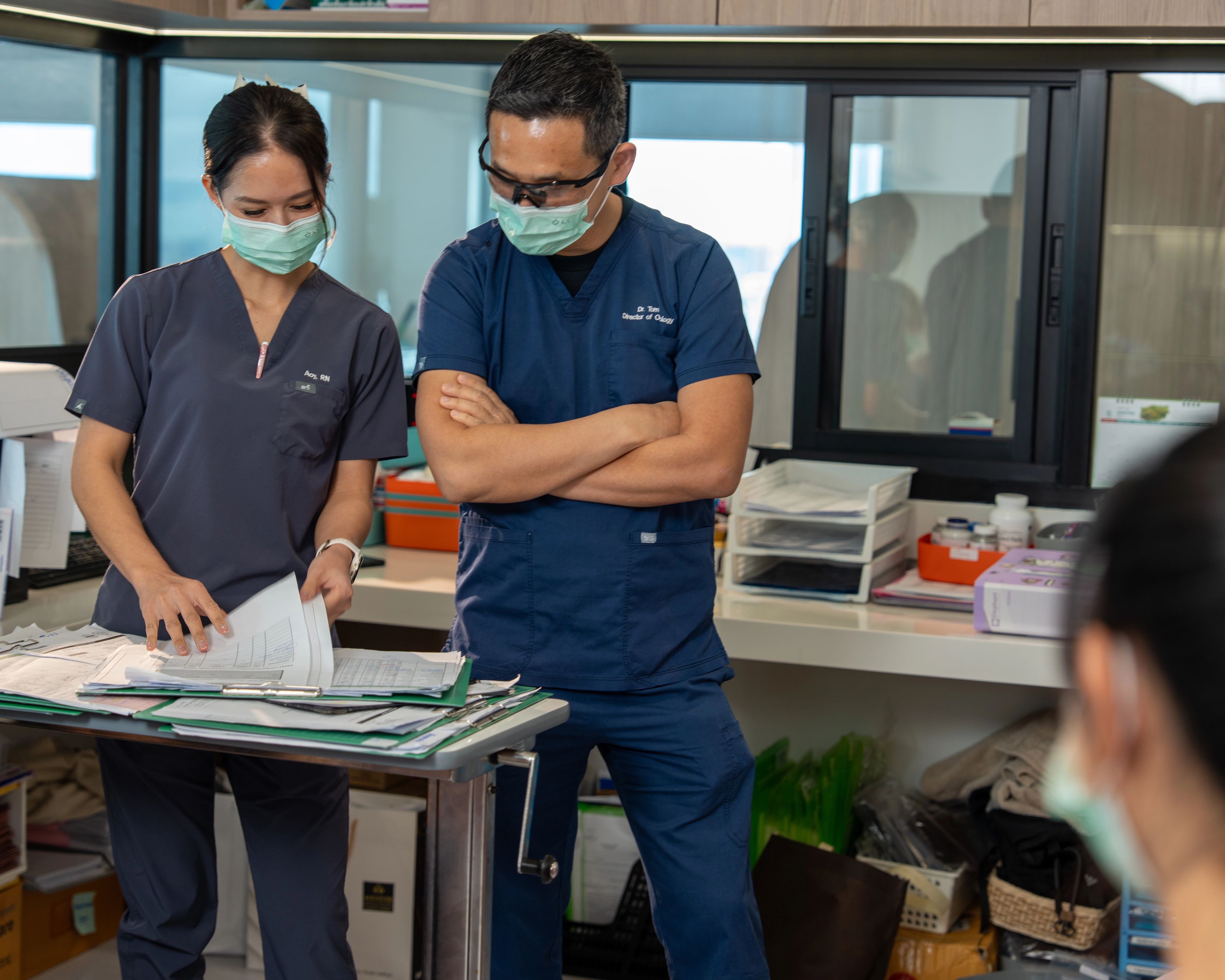 Nurse and doctor in scrubs and face masks having a conversation while looking at notes on a desk in an office, there is a third medical professional in the room.